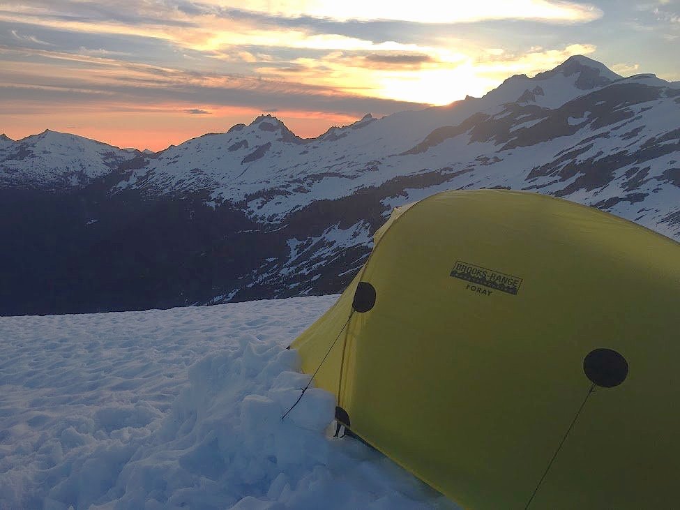 A yellow tent pitched on snowy terrain with snow-capped mountains and a colorful sunset sky in the background.