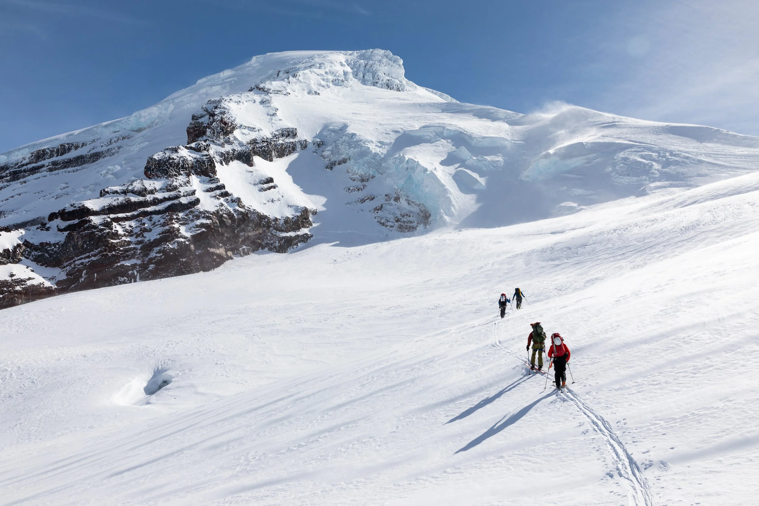 Group of four mountaineers climbing a snowy mountain slope under clear blue sky.