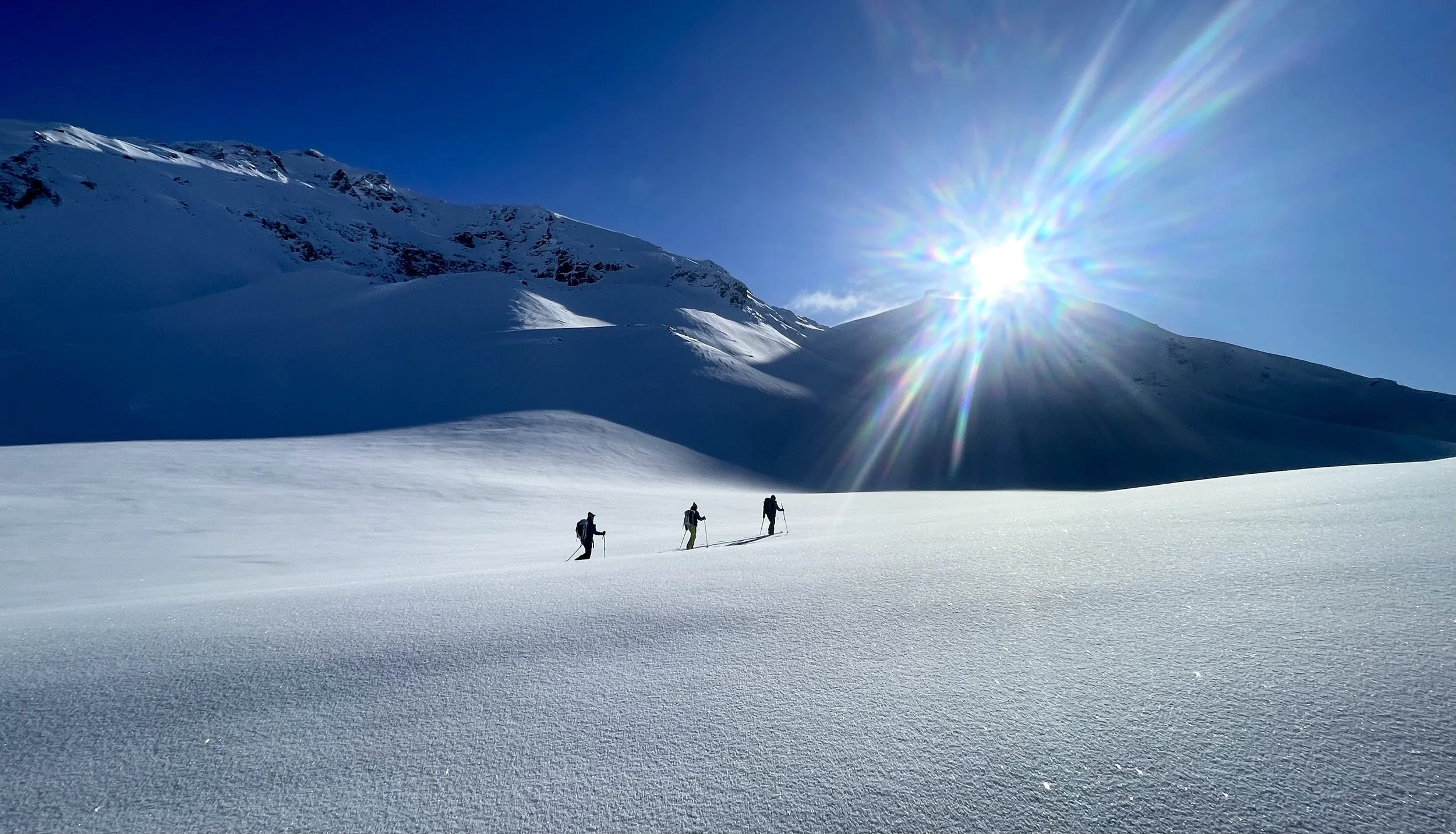 Three people hiking in a snow-covered mountain landscape with the sun shining brightly in a clear blue sky.
