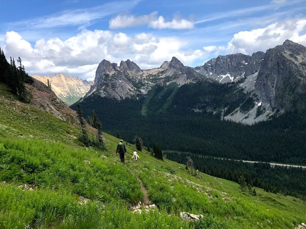 Hikers walking along a trail in a lush green mountain valley with towering rocky peaks in the background, partly cloudy sky above.