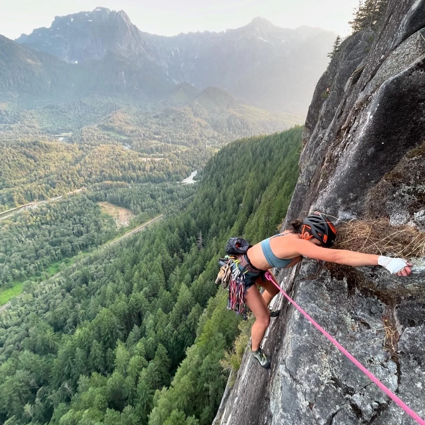 A woman rock climbing on a steep cliff with a scenic mountain and forest landscape in the background, wearing a helmet, climbing gear, and a sports bra.