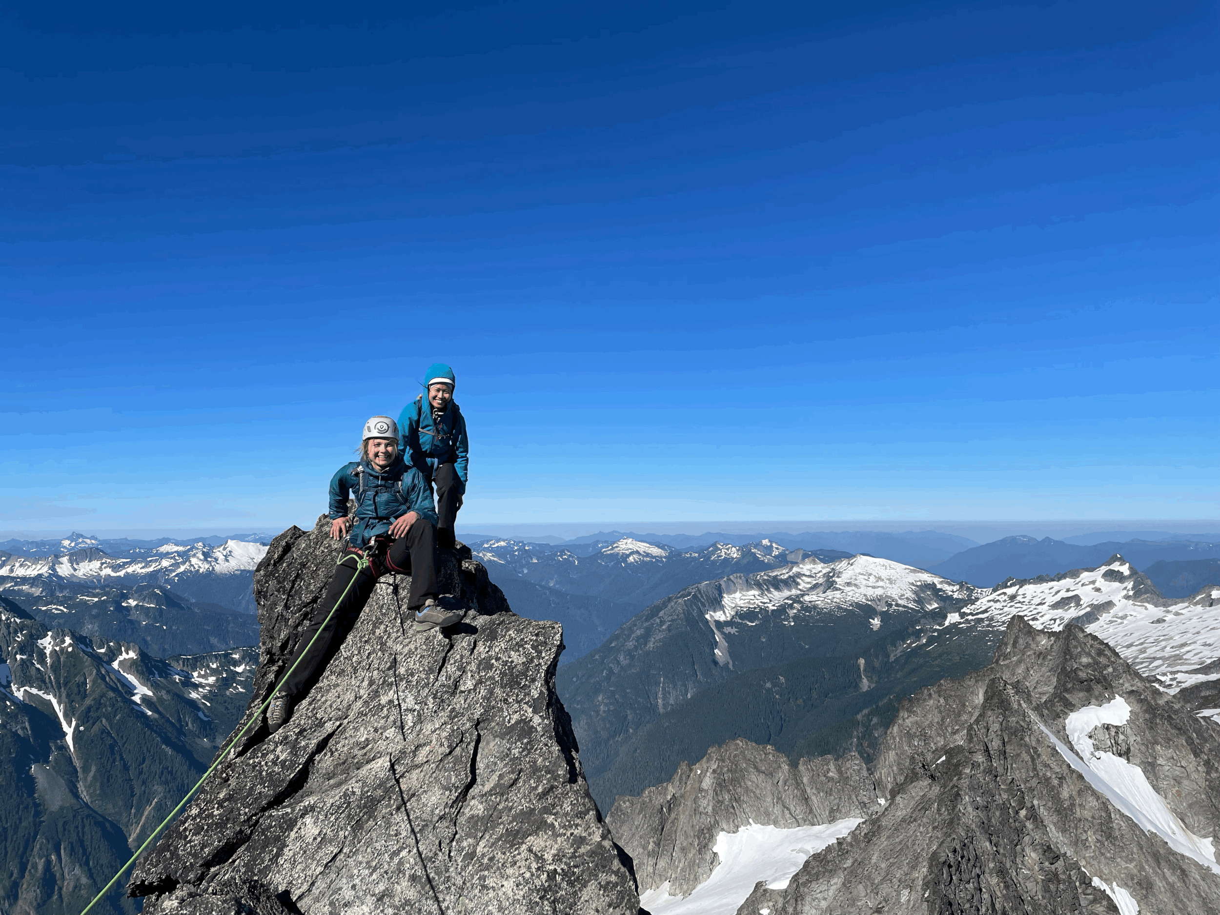 Two hikers wearing blue jackets and helmets sitting on a rocky mountain peak with snow-capped mountains in the background under a clear blue sky.