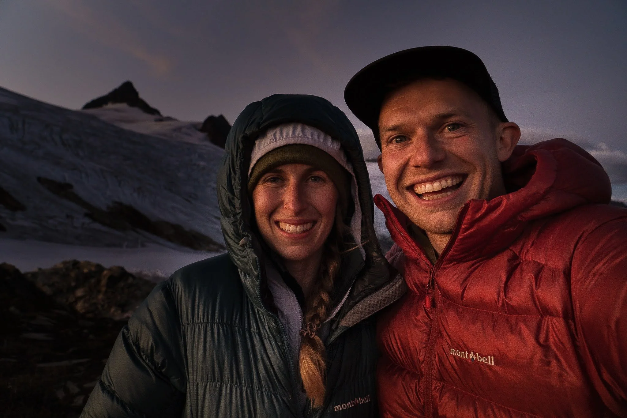 Two smiling hikers taking a selfie outdoors in a mountainous, snowy landscape at dusk, wearing outdoor jackets and hats.