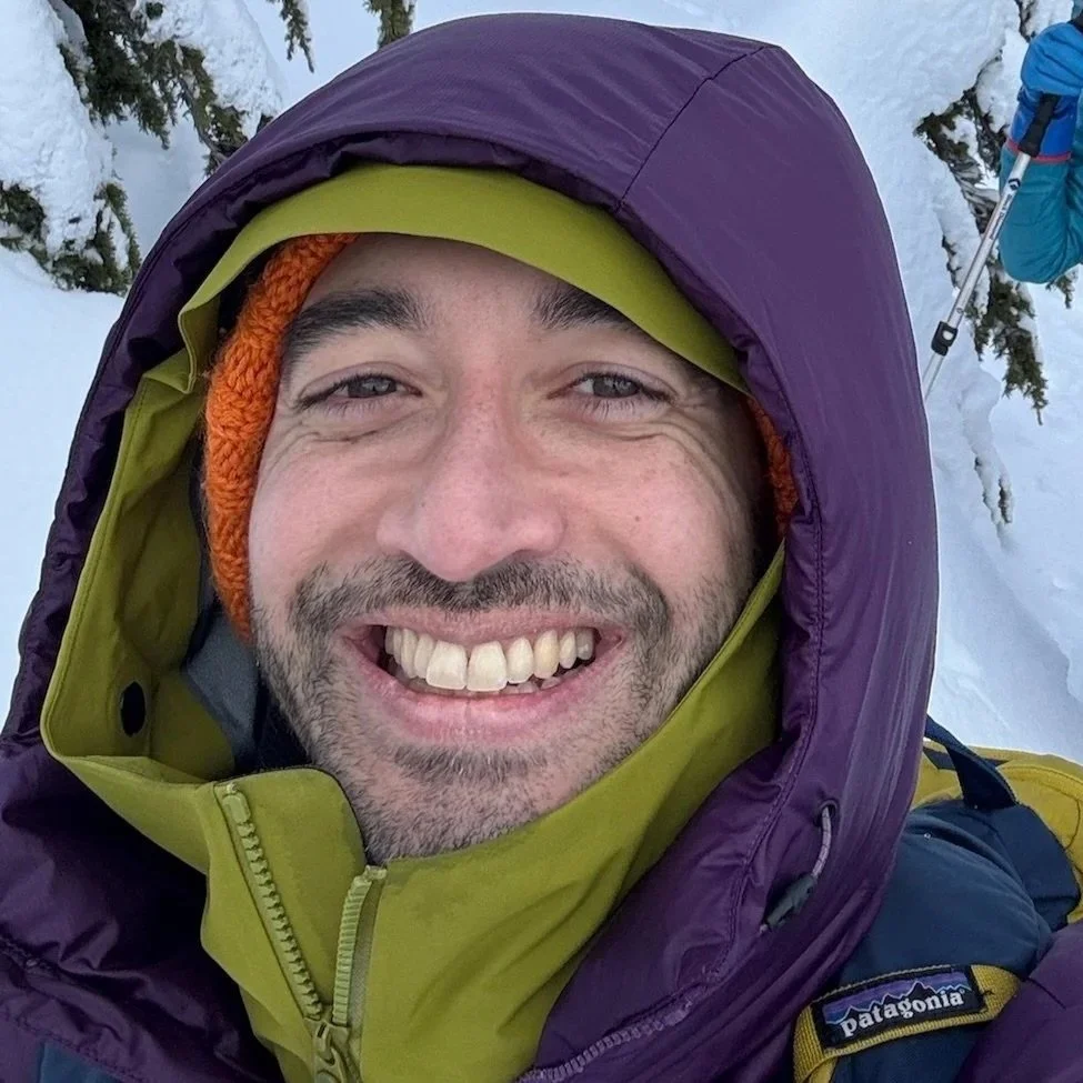 A smiling man taking a selfie in a snowy outdoor setting, dressed in winter gear with a Patagonia jacket, a purple hood, an orange knit hat, and surrounded by snow-covered trees.