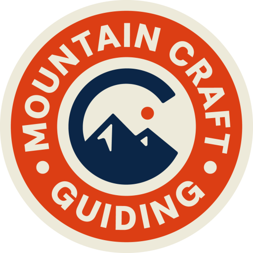 Circular logo with a red border and white text reading "Mountain Craft Guiding." Inside, a stylized mountain with a sun or moon overhead, all in navy blue and red.