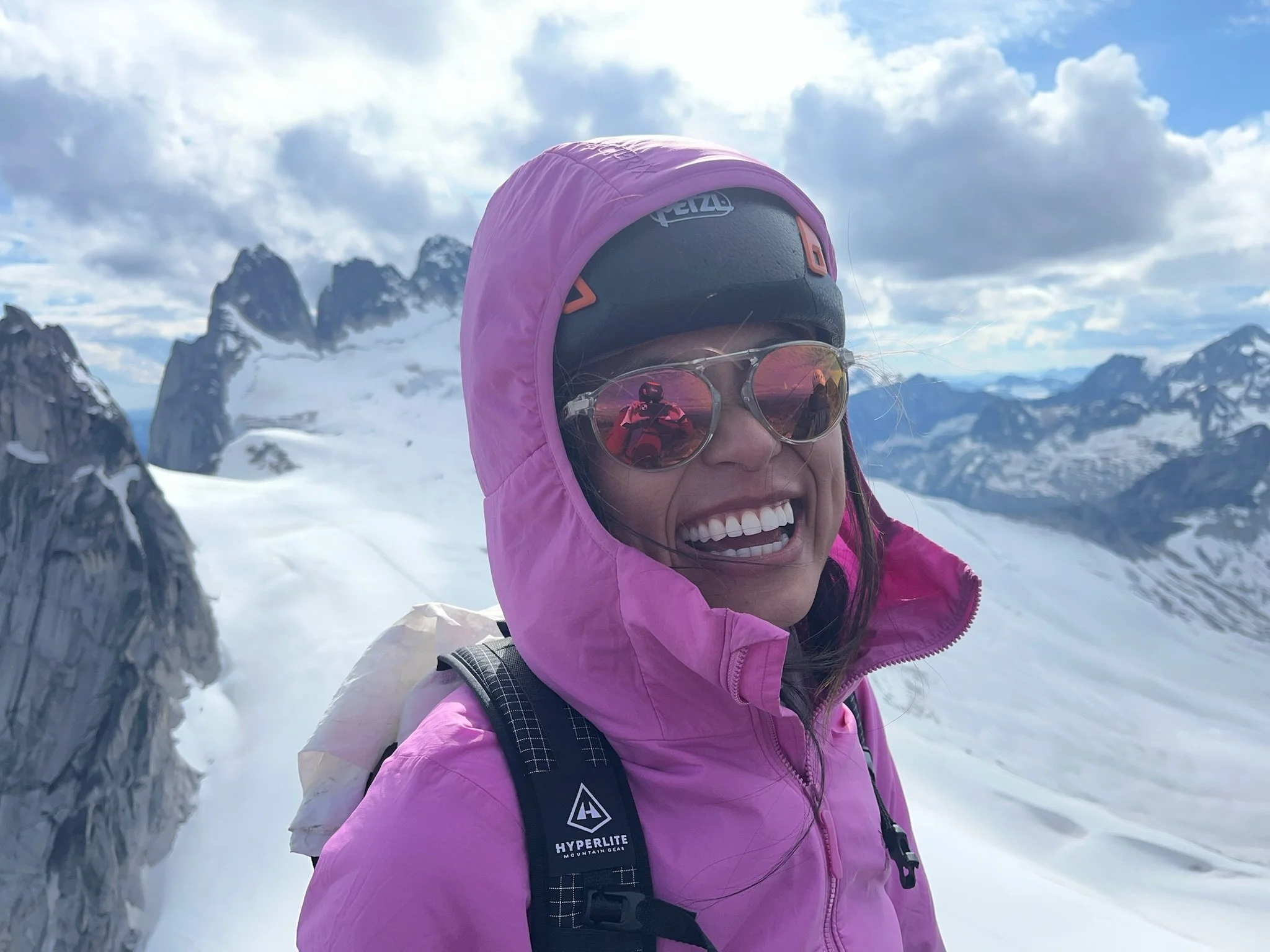 A woman in pink winter gear with sunglasses and a helmet, smiling on a snowy mountain landscape with rugged peaks in the background.