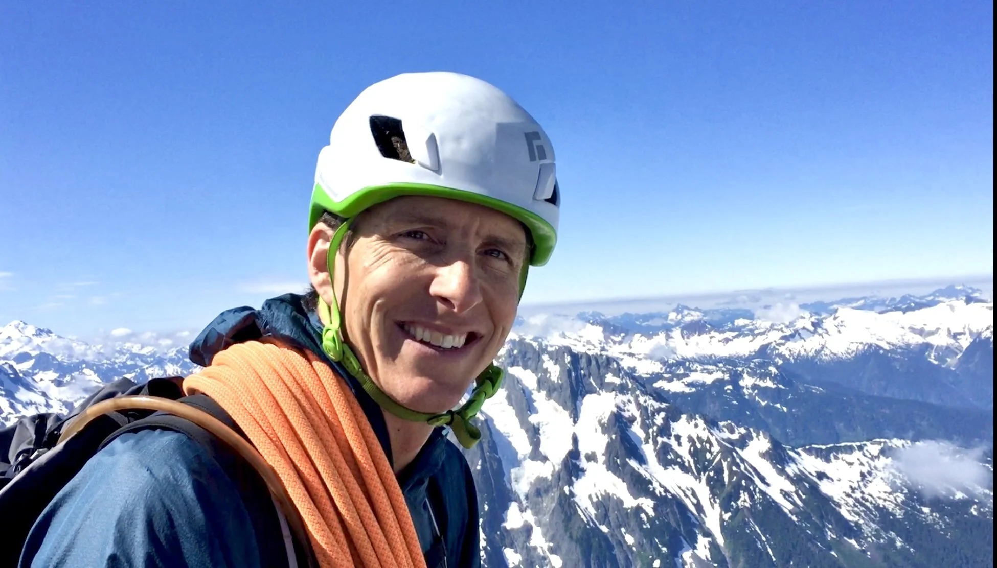 Man wearing a white and green helmet with a wide smile, standing on snow-covered mountain peaks under a clear blue sky.