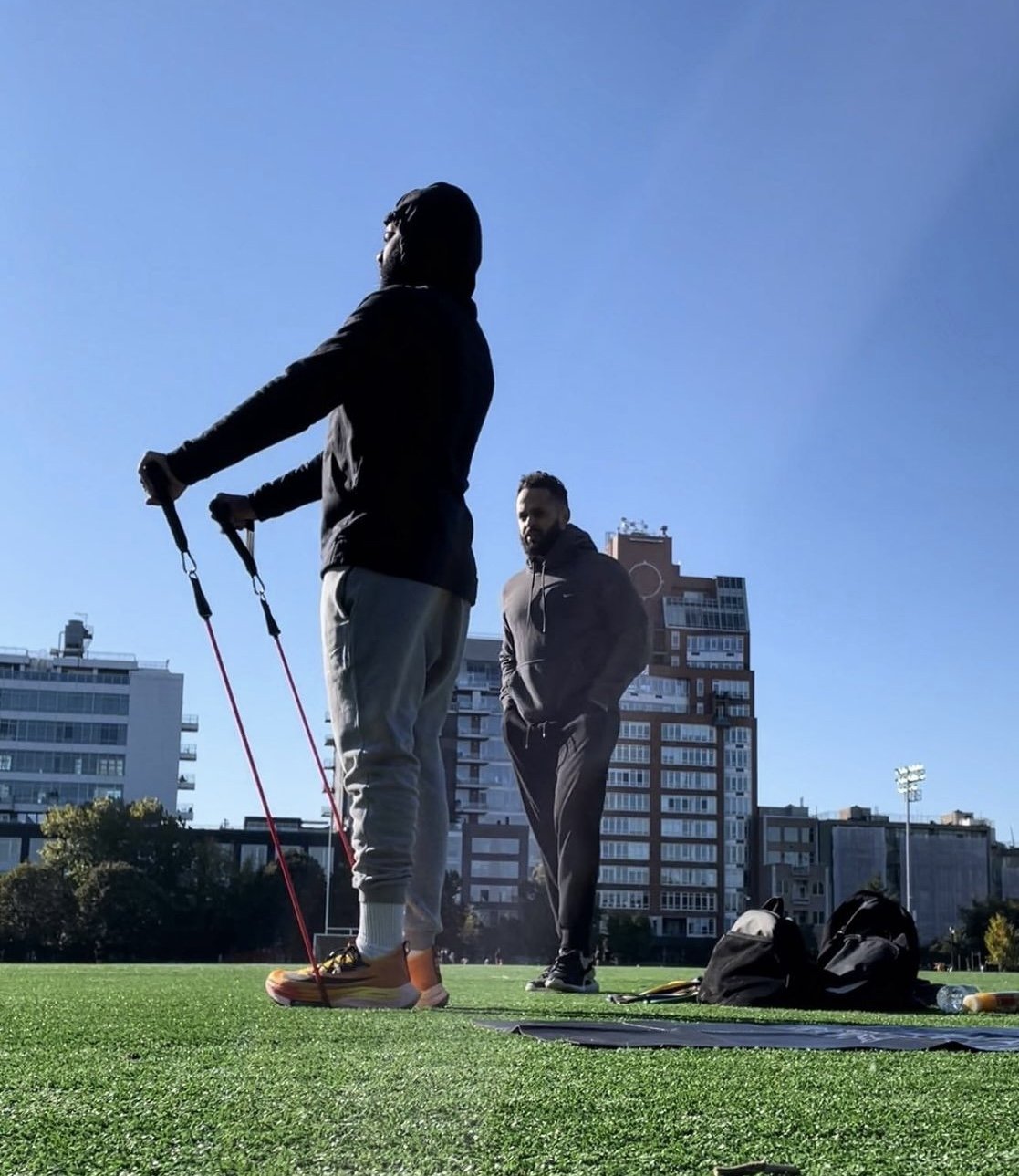 Two men standing on a grass field with buildings in the background. One man is holding exercise resistance bands, and the other is observing. There are bags and water bottles on the ground.