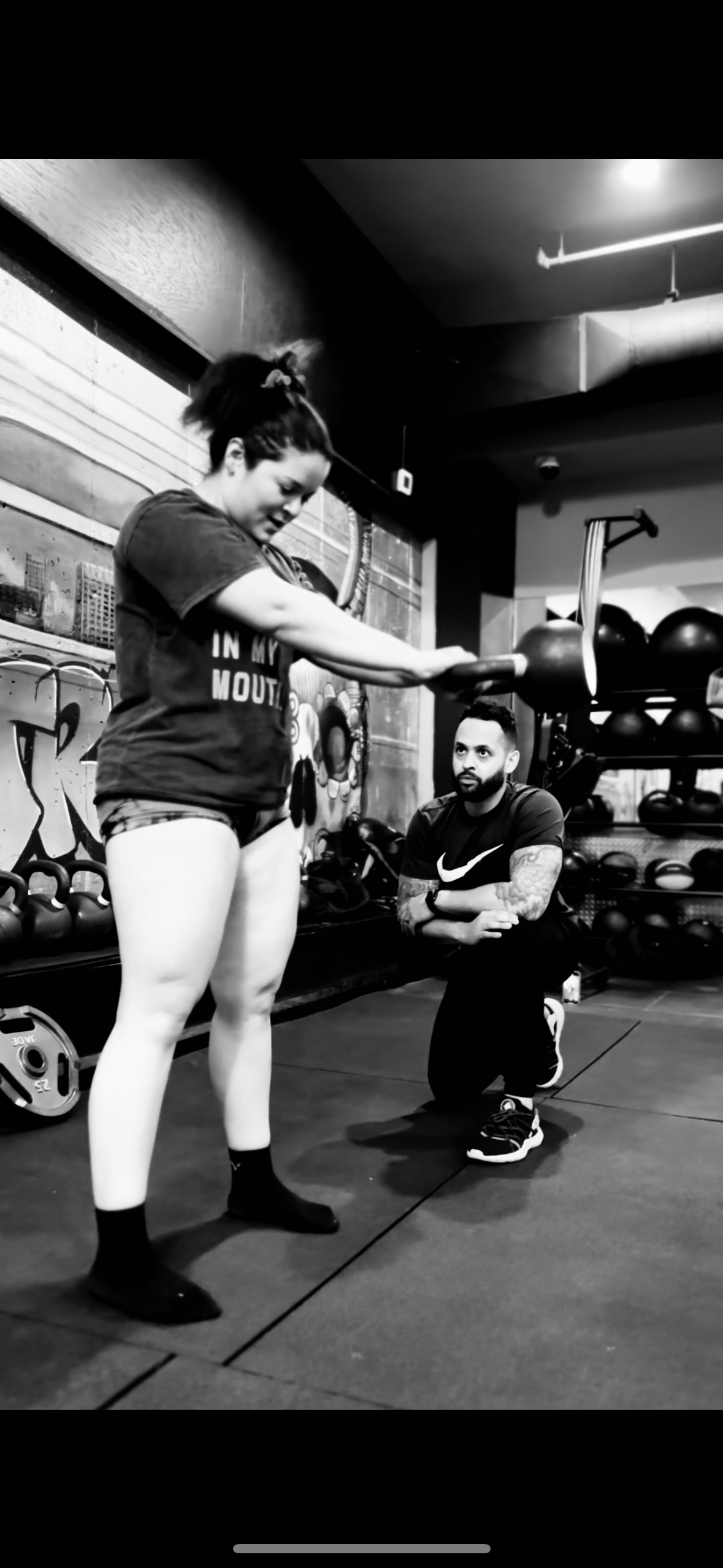A woman in workout clothes is weightlifting a barbell with weights on a squat rack, while a man kneels beside her, observing at a gym with racks of weights and gym equipment in the background.