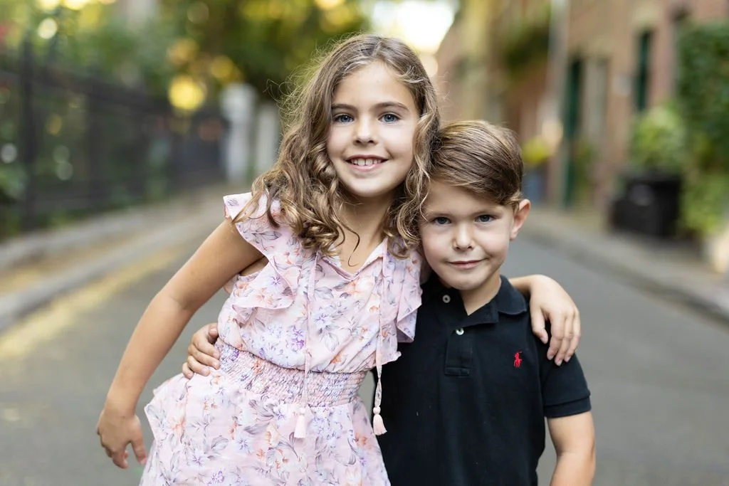 Two children standing closely together on a street with one arm around each other, one is wearing a floral dress and the other a black shirt.