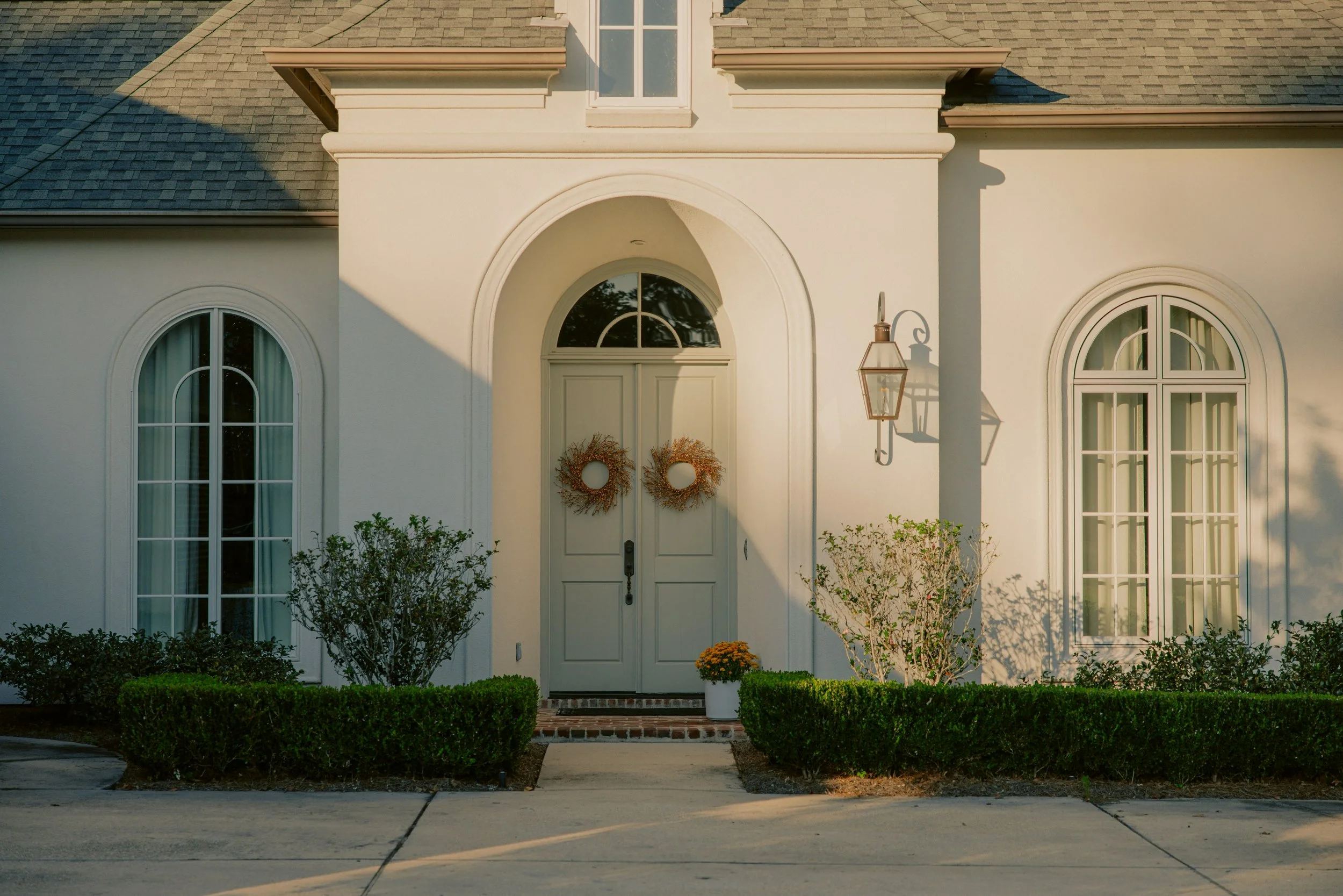 Front view of a white house with arched windows and a double door decorated with holiday wreaths, a brick step, potted plant, shrubs, and a large outdoor lantern.