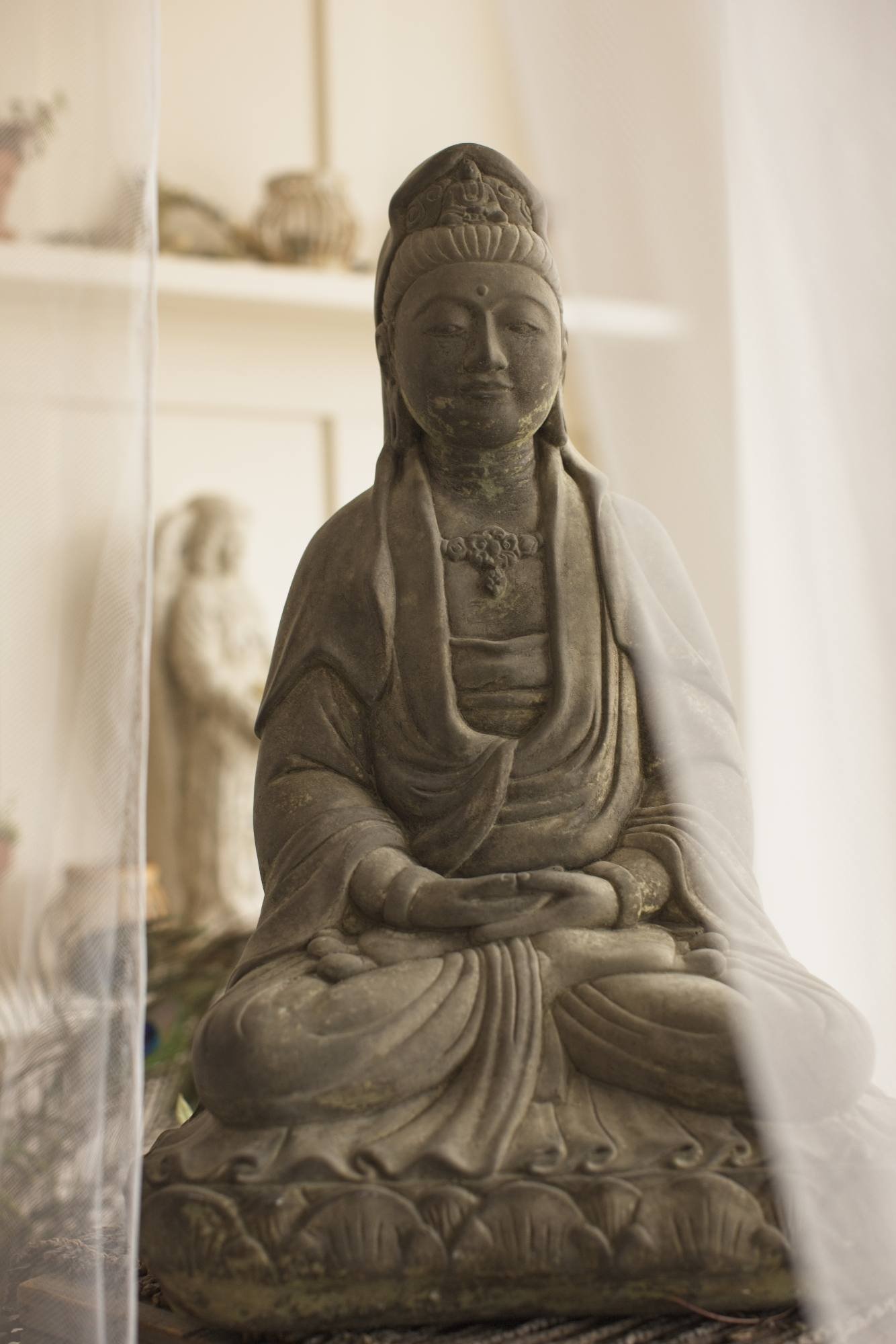 Photograph of a seated stone sculpture of a serene, smiling figure with traditional attire, adorned with a headdress, displayed indoors.