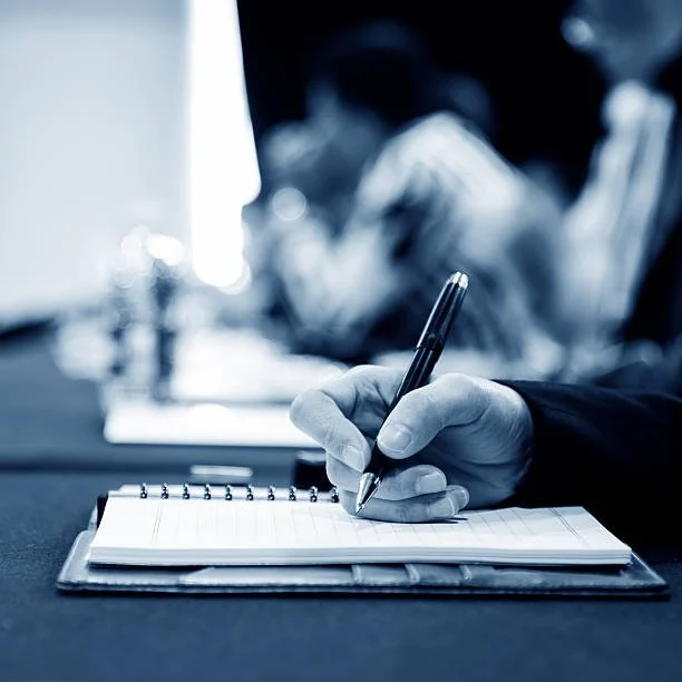Close-up of a hand writing with a pen in a notebook, with blurred background of people at a meeting or conference.