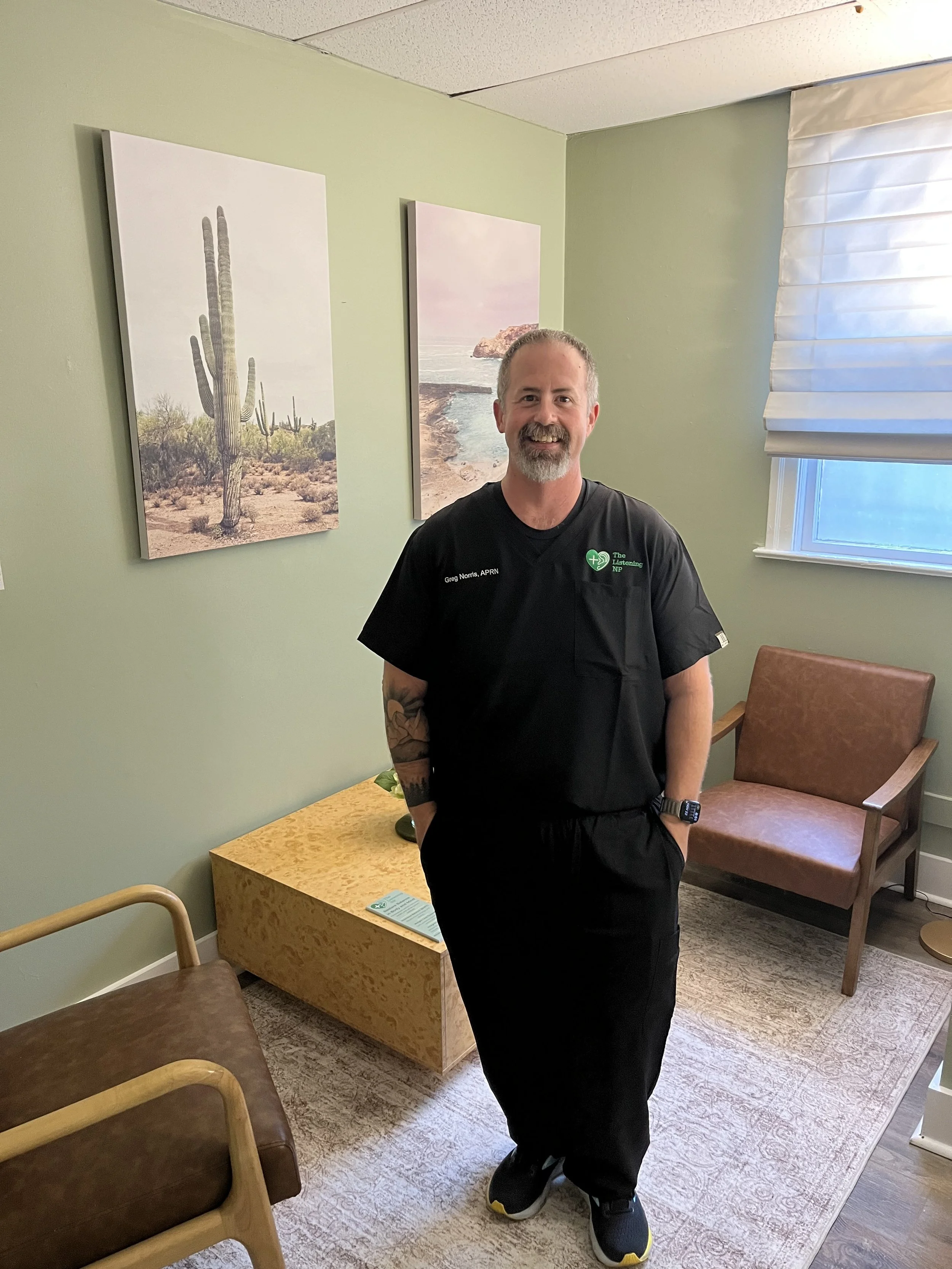 The Listening NP in black medical scrubs standing in a room with light green walls and desert-themed artwork, near a window with white blinds and a brown armchair.