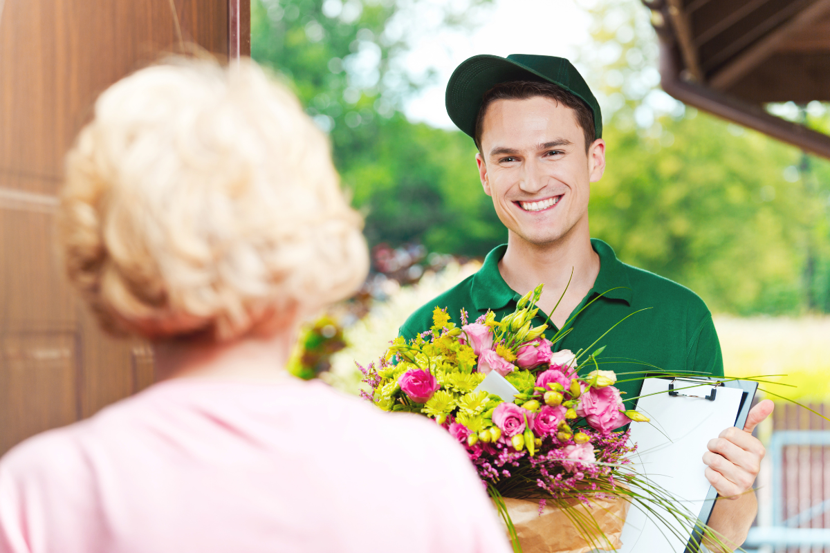 A Gifts of Flowers designed by TA located in Agawam MA delivery man smiling while holding a large bouquet of pink and yellow flowers. Delivers flower to an older woman wearing a pink top is facing him.