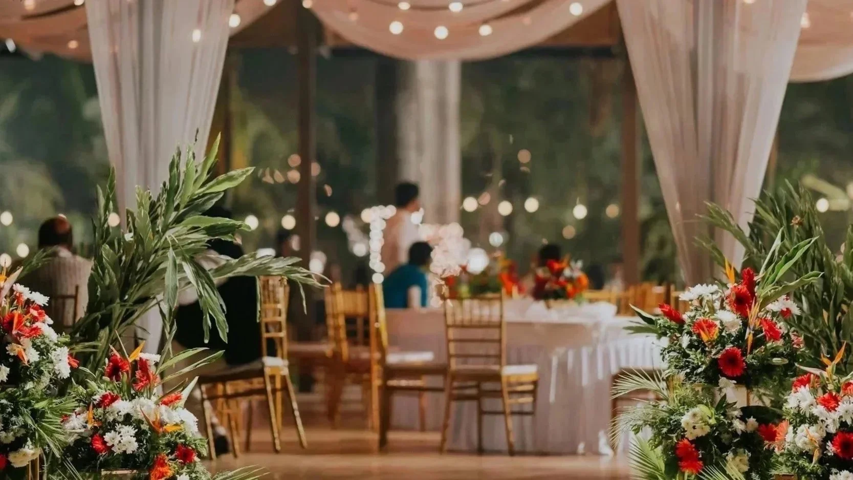 Decorated event space with floral arrangements, round tables, and chairs, set for a celebration or wedding reception, with blurred background of people and twinkling lights.