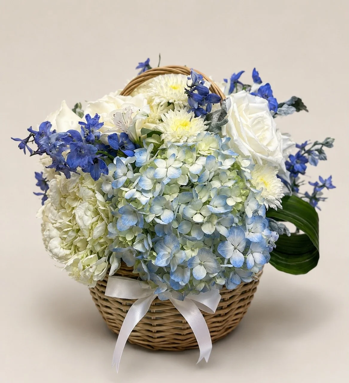 A wicker basket filled with white and blue flowers, including hydrangeas, roses, and other small blossoms, decorated with a white ribbon.