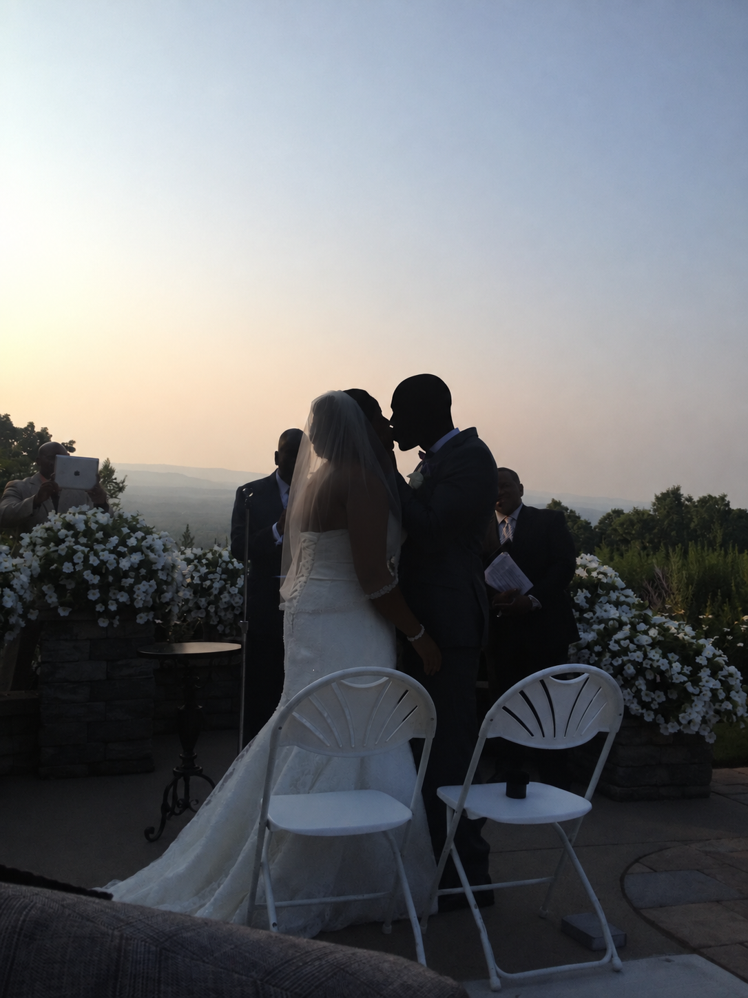 A silhouette of a bride and groom kissing during their outdoor wedding ceremony at sunset, with floral arrangements and guests in the background.