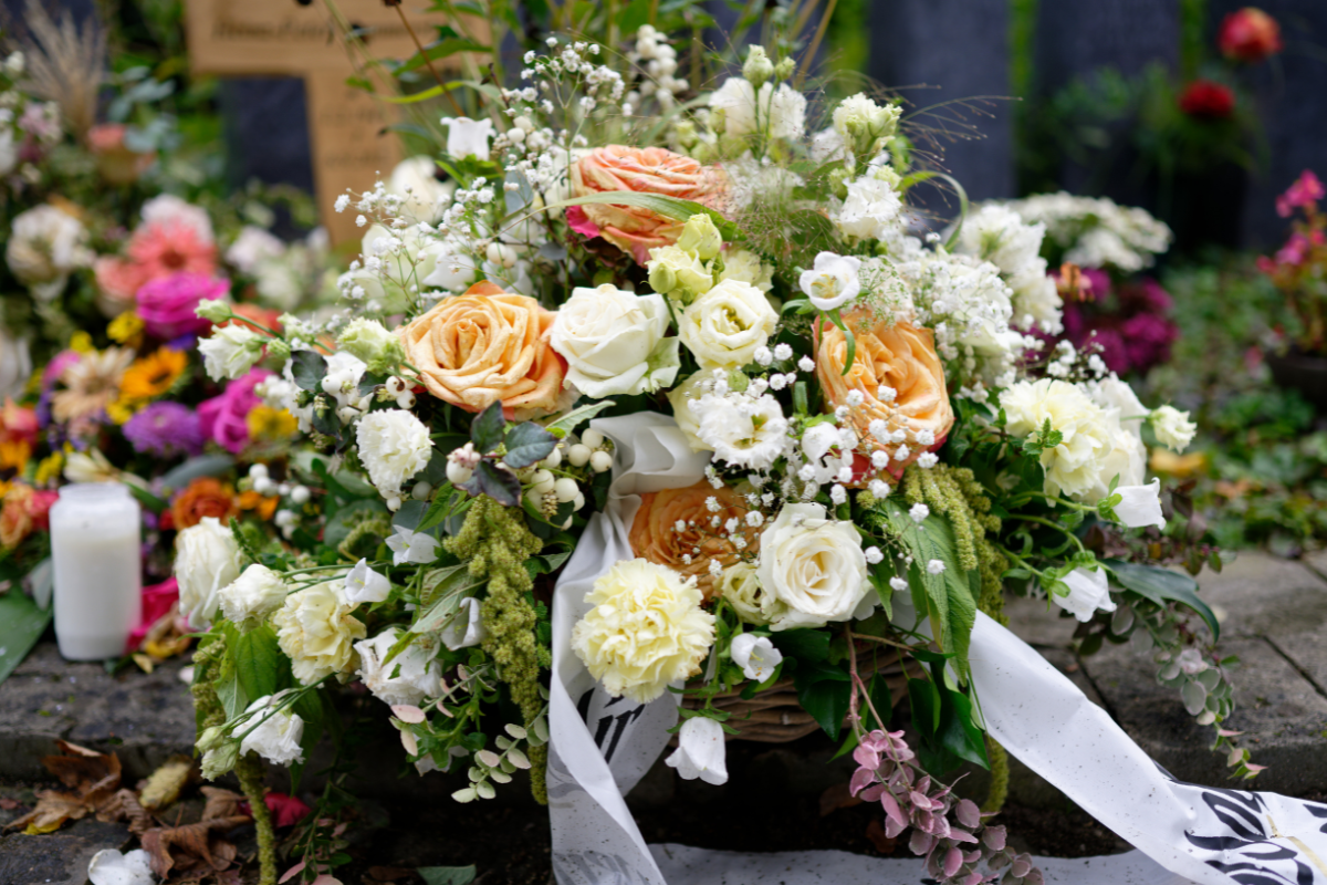 A floral wreath with white, cream, and peach roses, white baby's breath, and greenery, placed on a stone surface at a memorial or funeral ceremony, designed with intent by Gifts of Flowers designed by TA to as Tribute to A Loved One
