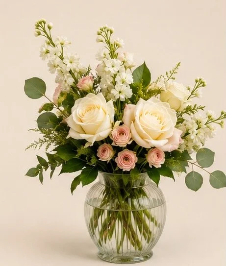 A glass vase with white and pink roses, white stock, and greenery on a white background.