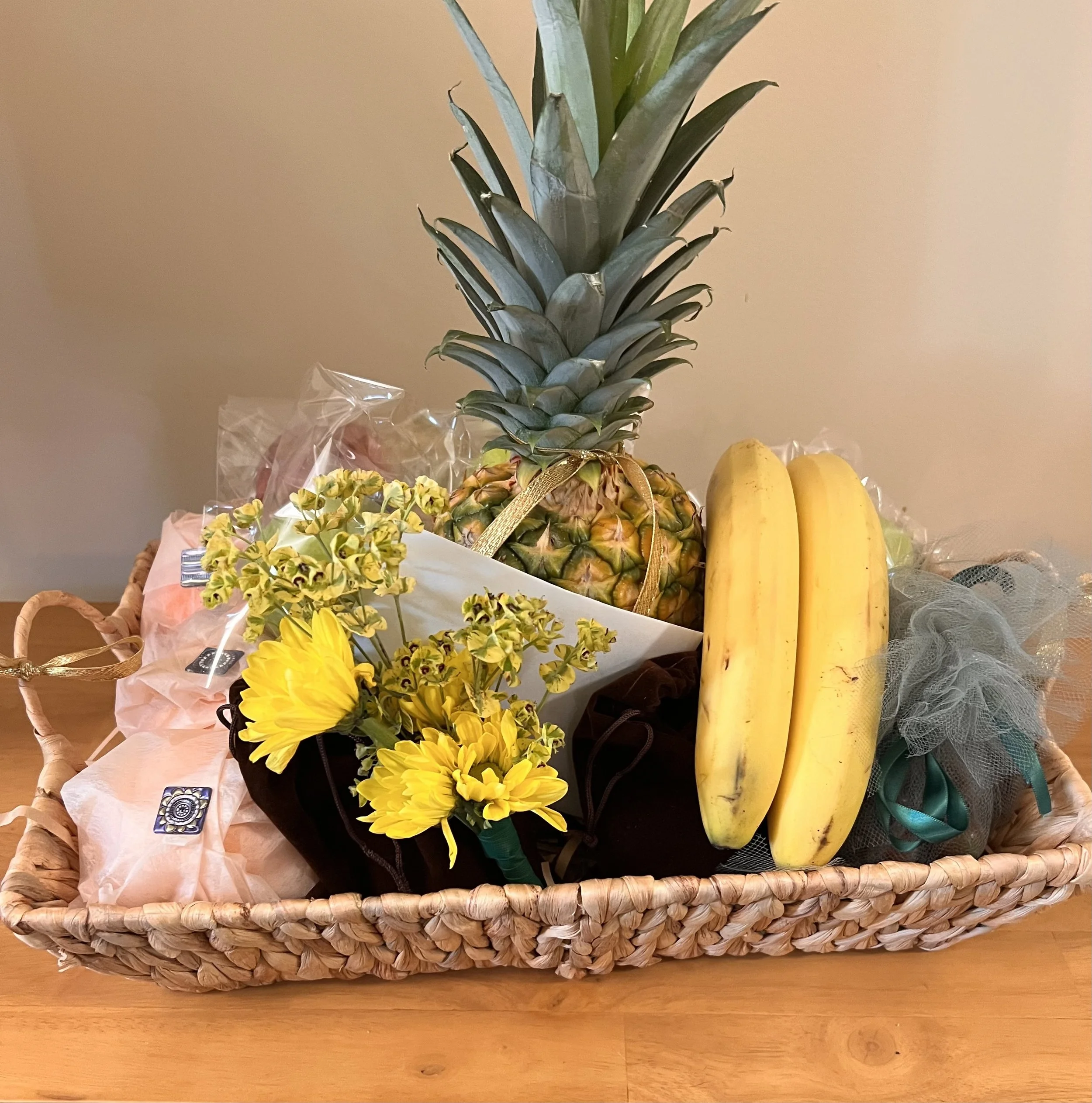 Fresh pineapple with green leaves on top, two bananas, and yellow flowers in a black basket on a wooden surface. fruit baskets delivered locally to residents and nursing -rehab facilities
