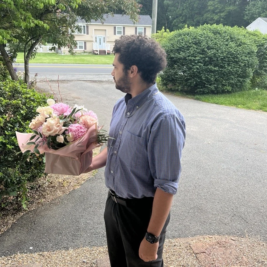 Alexavier wearing a checkered blue shirt and black pants stands outdoors, holding a bouquet of pink and white flowers wrapped in pink paper, with a background of trees, bushes, and a house.