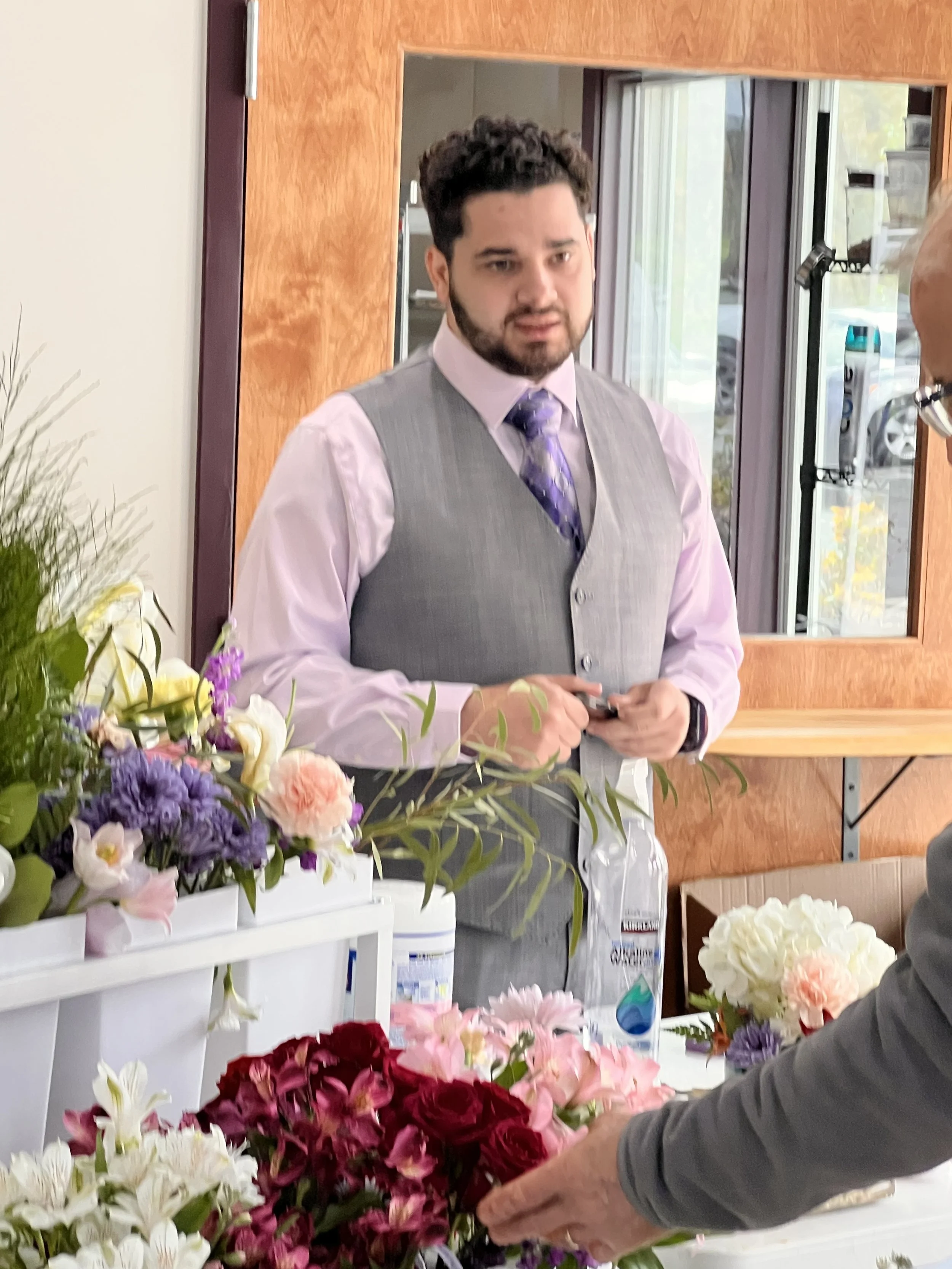Alexavier in a gray vest and purple dress shirt with a purple tie, standing at a floral display, talking to a customer with their hand on a bouquet of pink and red flowers, inside a flower shop.