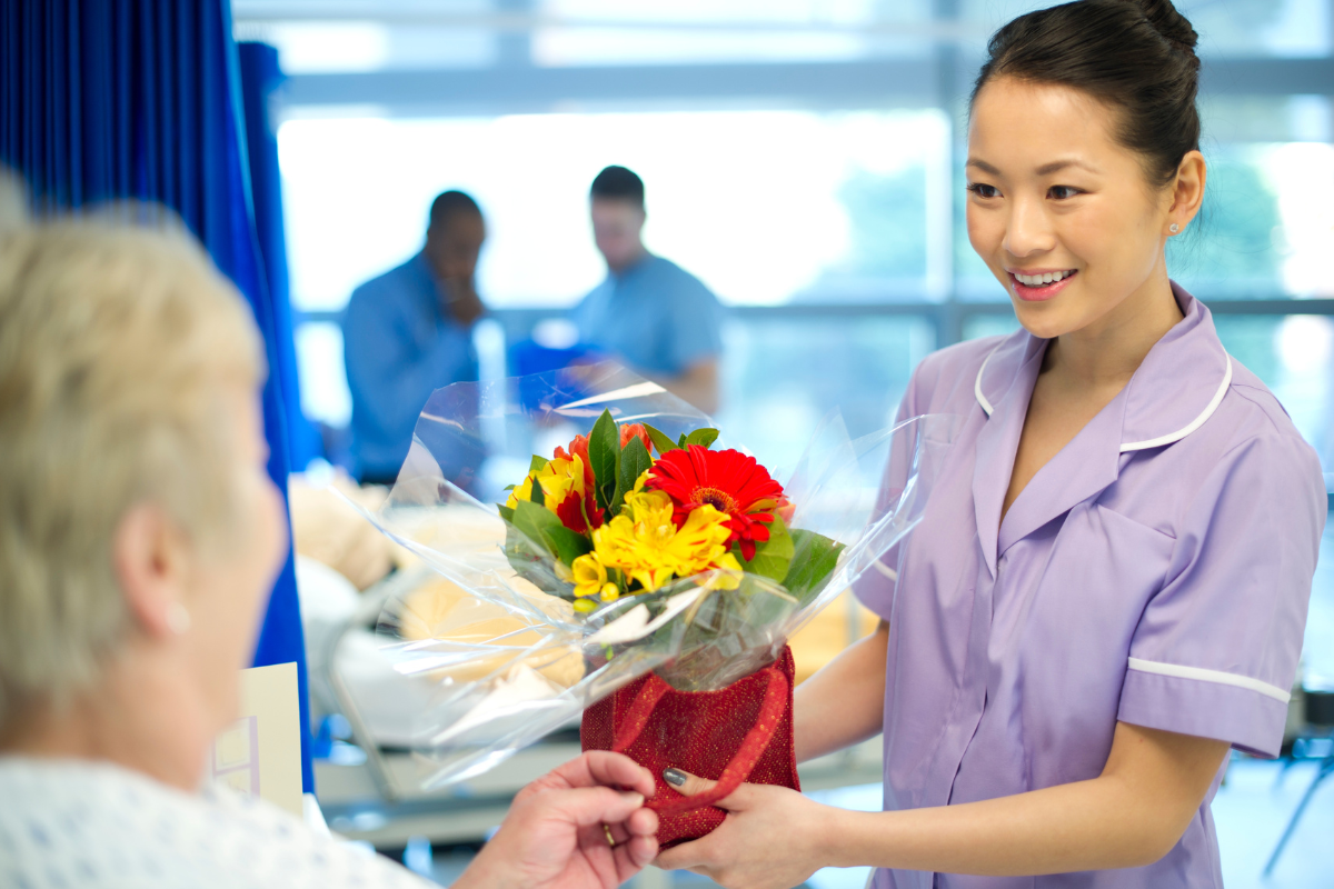 A young woman in lavender uniform giving a colorful bouquet of flowers Delivered by Gifts of Flowers designed by TA to an older woman in a hospital gown in a bright hospital room.