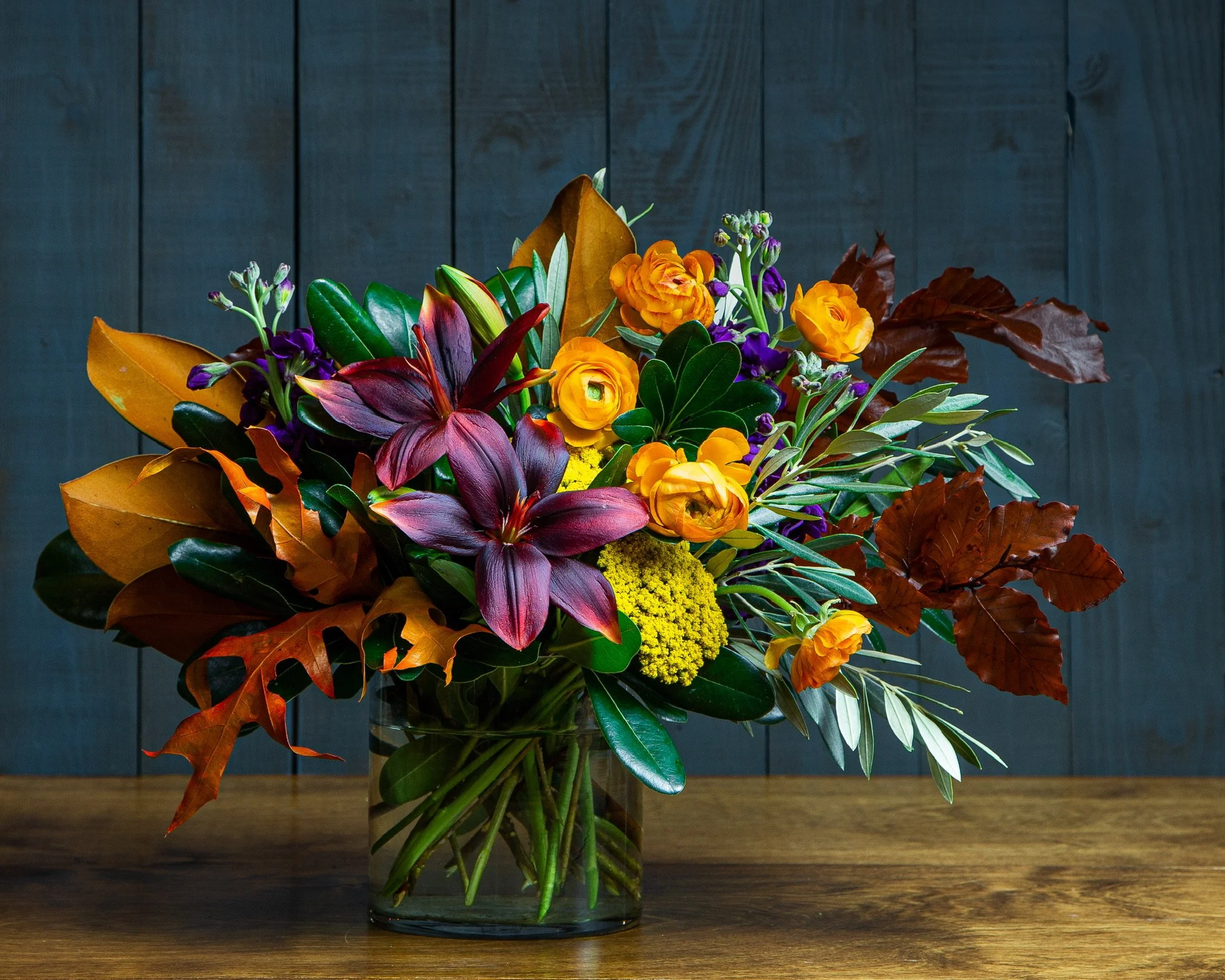 “Autumn bouquet featuring burgundy lilies, orange ranunculus, golden fillers blooms with hints of purple. accented with rustic oak leaves, dried magnolia leaves and luscious greens."
