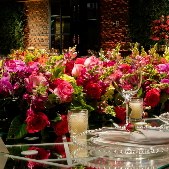 Elegant table setting with a large arrangement of pink and red roses and flowers, glassware, candles, and silverware, outdoors at night with brick building and greenery in the background.