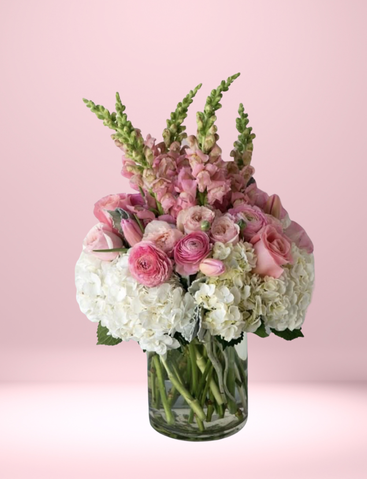 Soft blush and ivory floral arrangement in a clear glass vase featuring ranunculus, hydrangea, roses, snapdragons, and seasonal greenery.