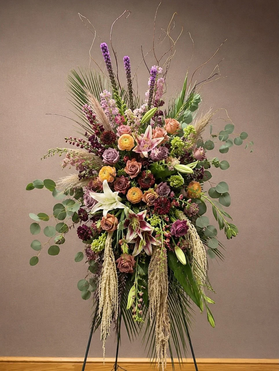 A large floral arrangement on a stand with pink, purple, white, and yellow flowers including lilies, roses, and other blooms, accented with green leaves and long, wispy branches.