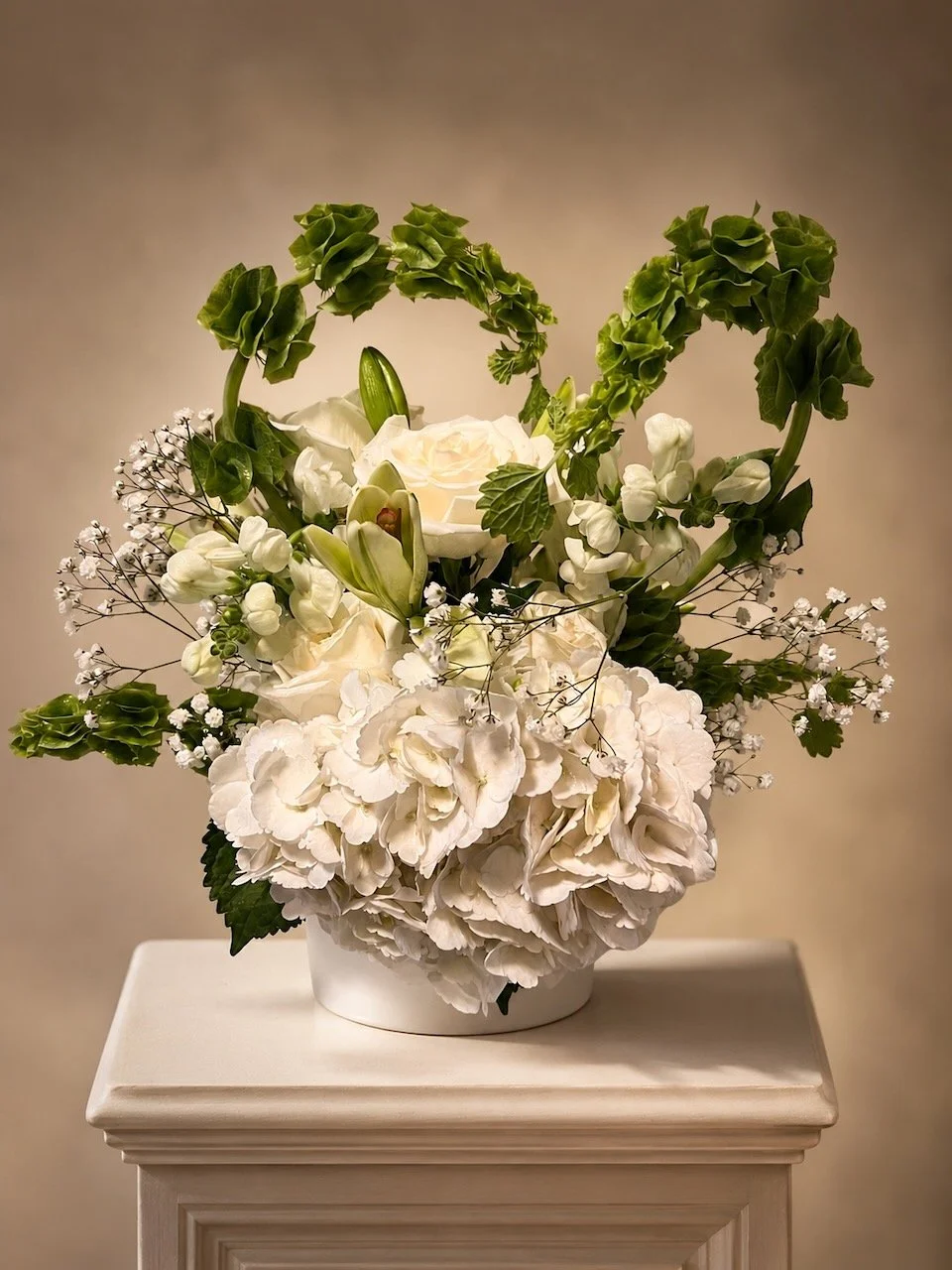 White floral arrangement with roses, hydrangeas, and greenery in a white vase on a white pedestal.