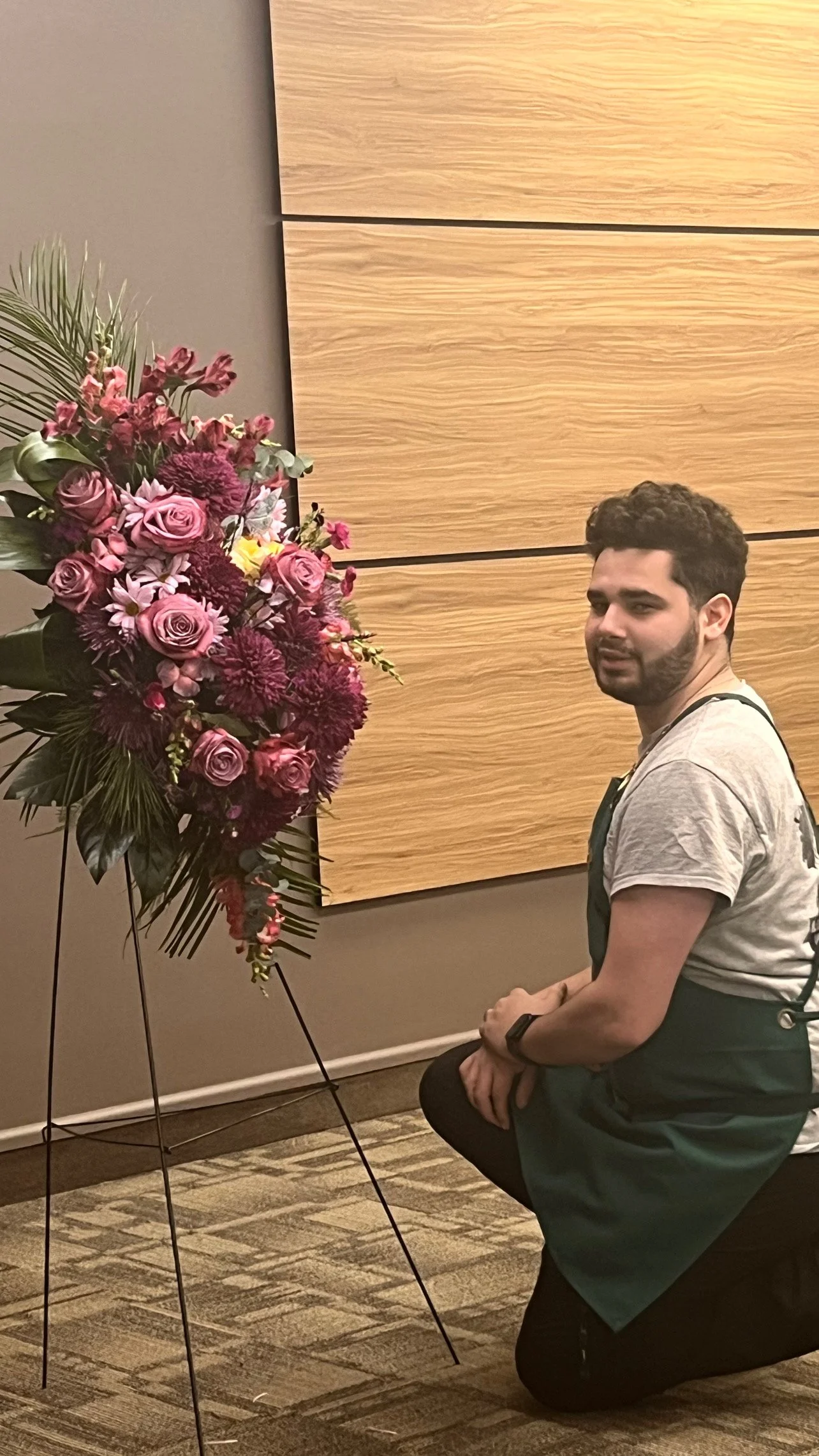Alexavier Davis  owner of Gifts of Flowers kneeling on a carpeted floor next to a large floral display with pink and purple flowers, against a wooden wall and gray background.