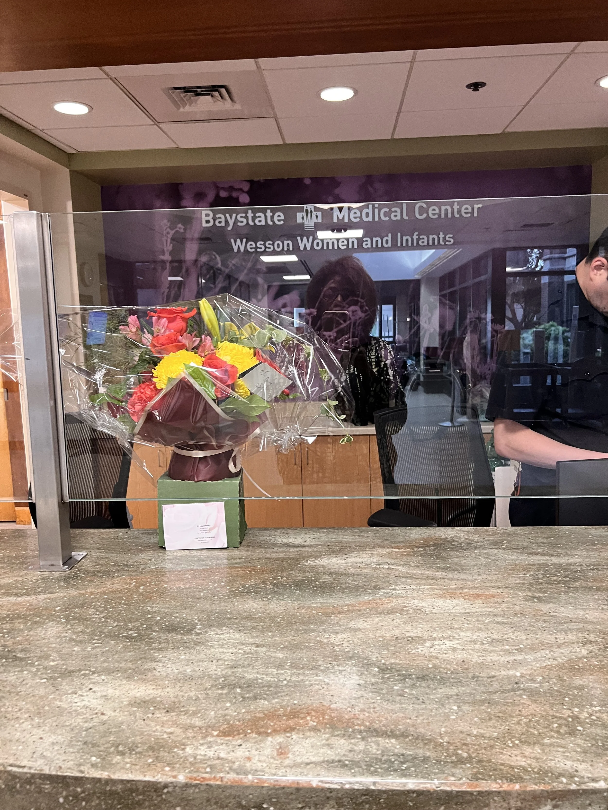 A floral bouquet with red, yellow, and pink flowers wrapped in plastic and placed on a counter in front of a glass window at the Baystate Medical Center, Wesson Women and Infants section.