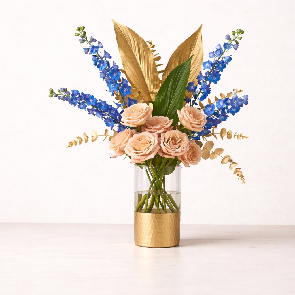 Modern garden luxe arrangement with champagne roses, blue delphinium, and fresh foliage with gold accents. Elegant and thoughtfully designed.