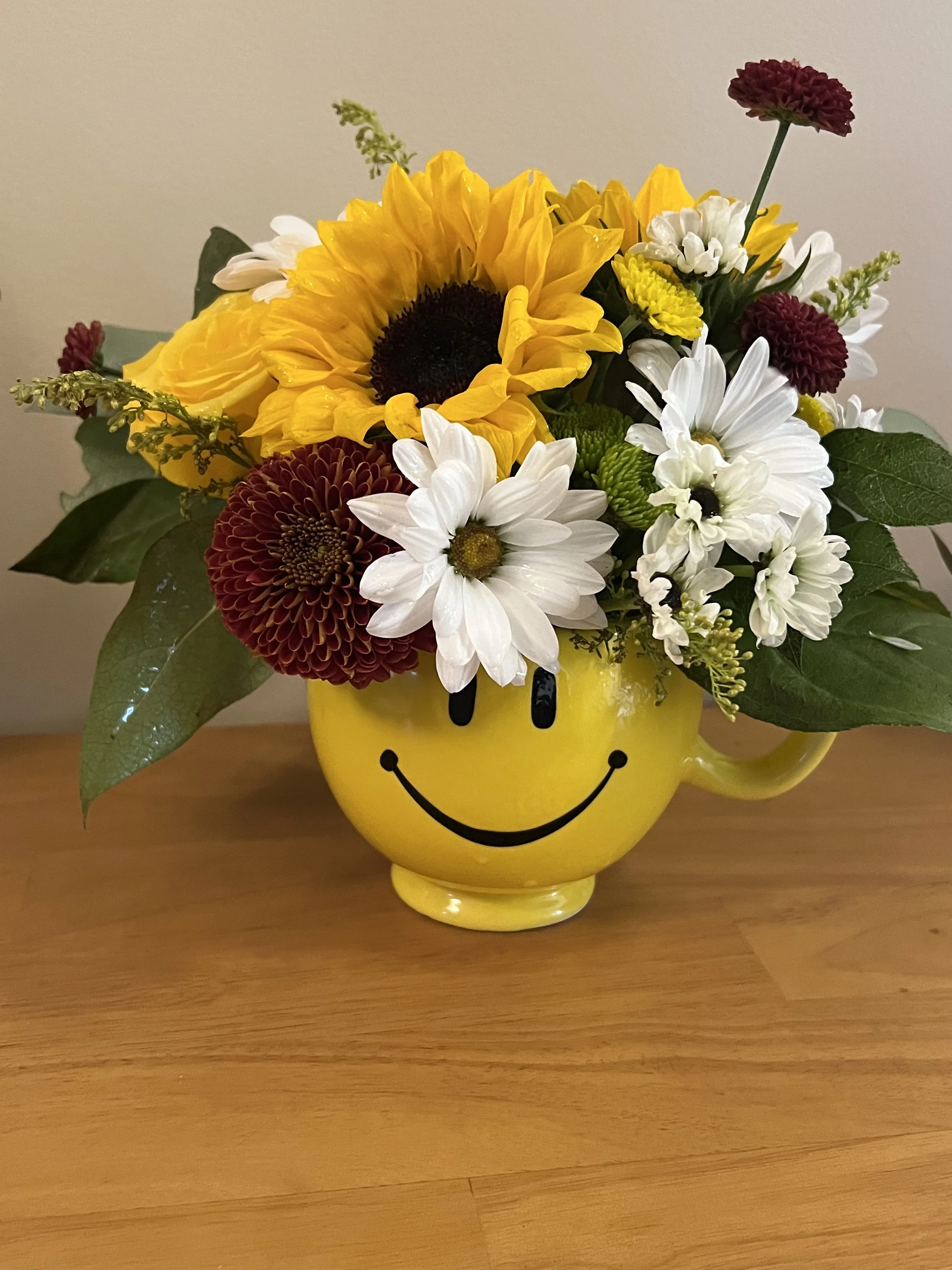 A yellow smiley face mug filled with a colorful bouquet of flowers, including sunflowers, daisies, coxcombs, and other assorted blooms, sitting on a wooden surface. Get well soon and happy office mugs