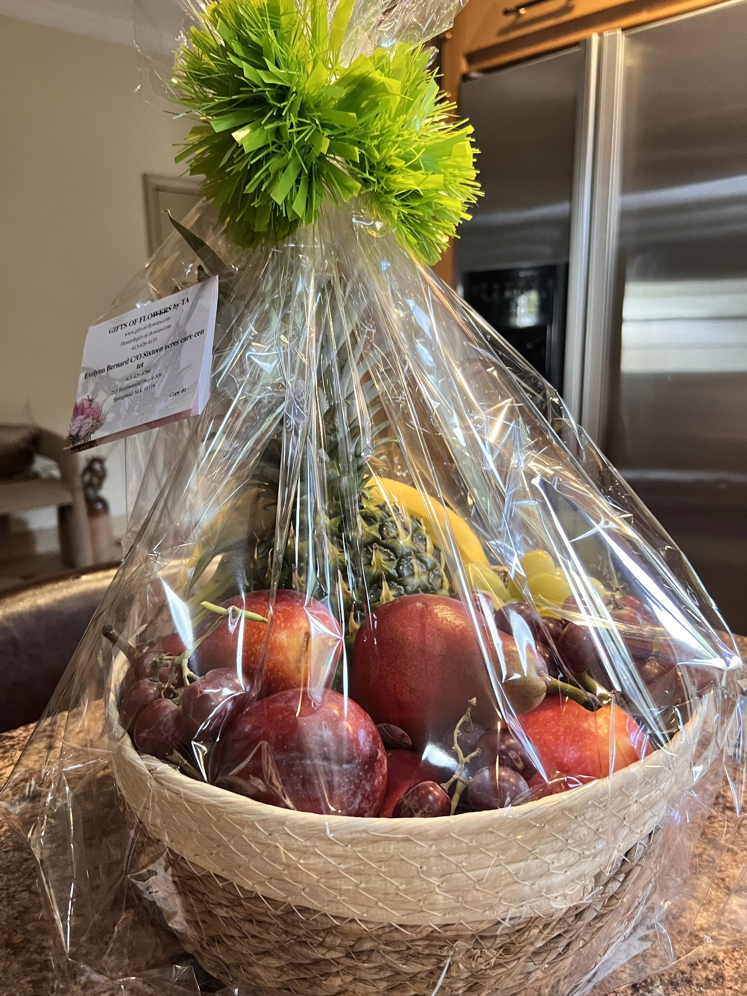 A basket of fresh fruits, including pineapples, apples, and grapes, wrapped in clear plastic wrap. Gifts of Flowers designed by TA delivers fruit baskets