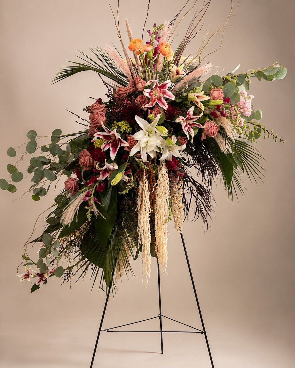 A large floral arrangement featuring pink roses, white lilies, pink and yellow ranunculus, eucalyptus leaves, and various greenery on a black metal stand.