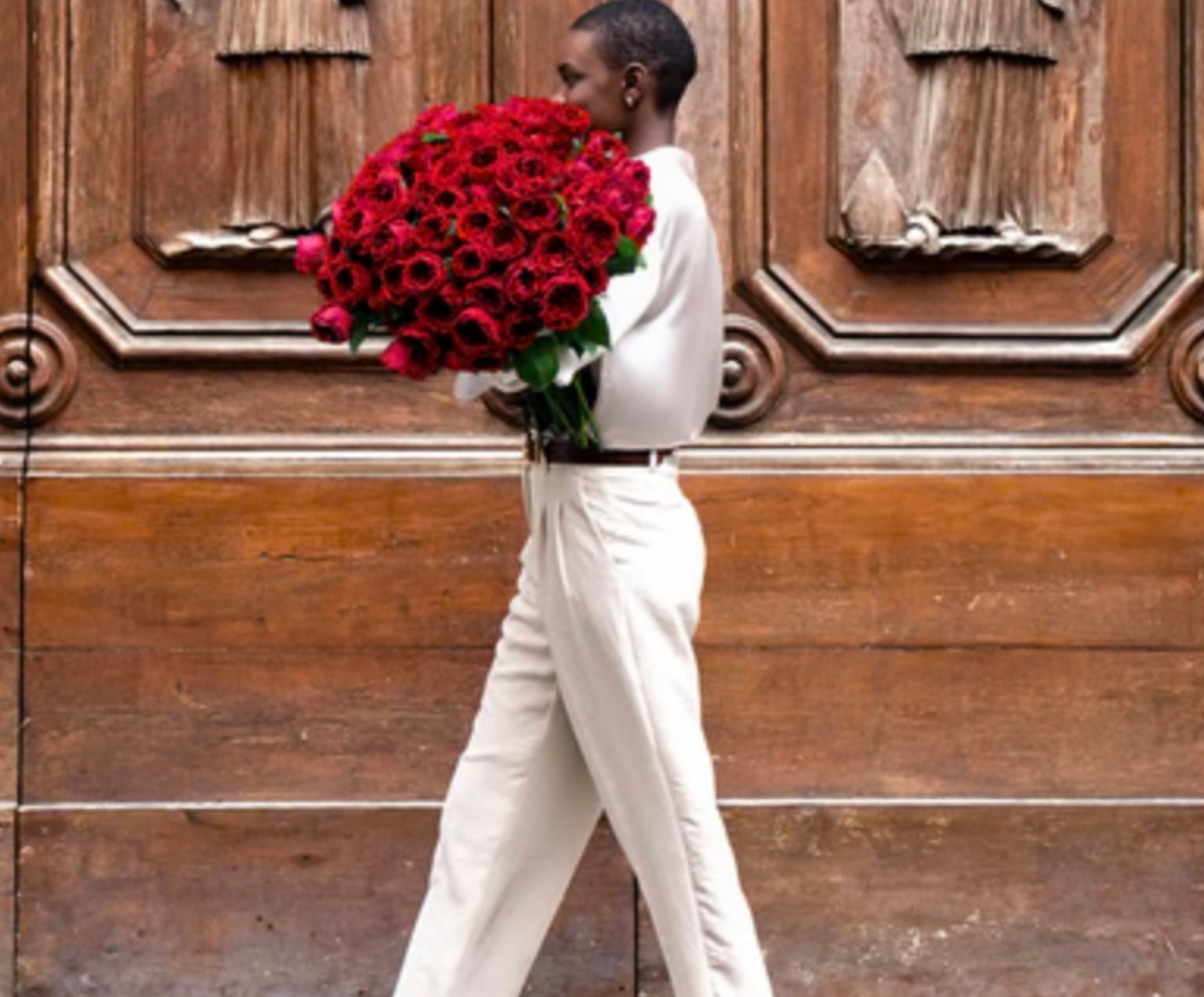 A woman with short hair and dark skin in white clothing carrying a large bouquet of red roses in front of a wooden door.