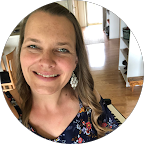 A woman with shoulder-length brown hair smiling at the camera, wearing earrings and a floral top, standing indoors in a well-lit room.