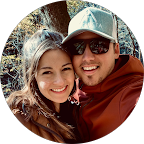 A smiling young woman and young man taking a selfie outdoors with a body of water and mountains in the background.