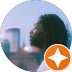 Side profile of a man with glasses and dreadlocks outdoors during sunset with city buildings in background