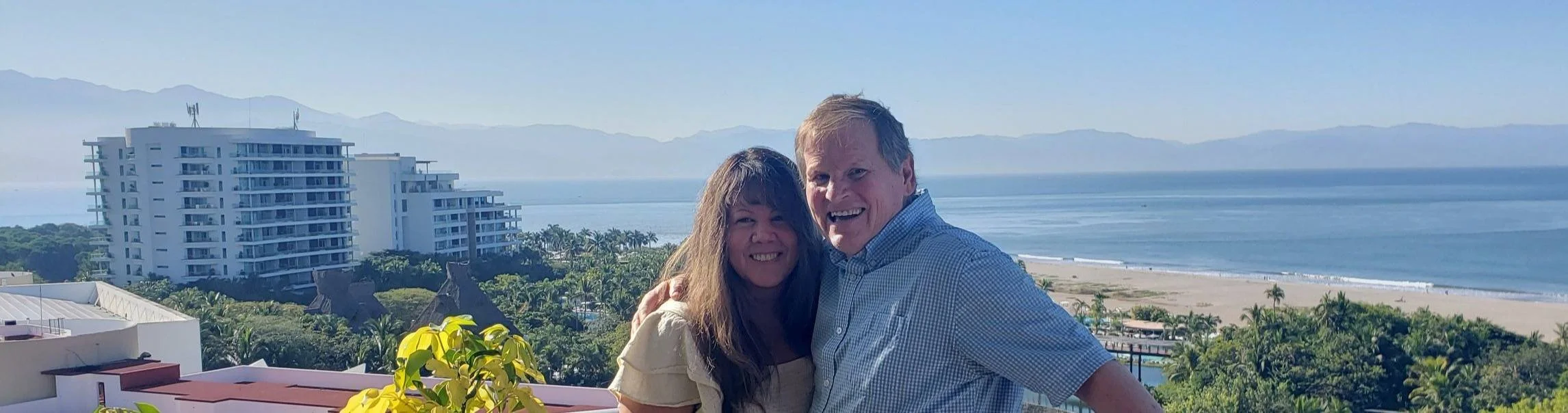 Smiling couple standing on a balcony with a beach, ocean, and mountains in the background.