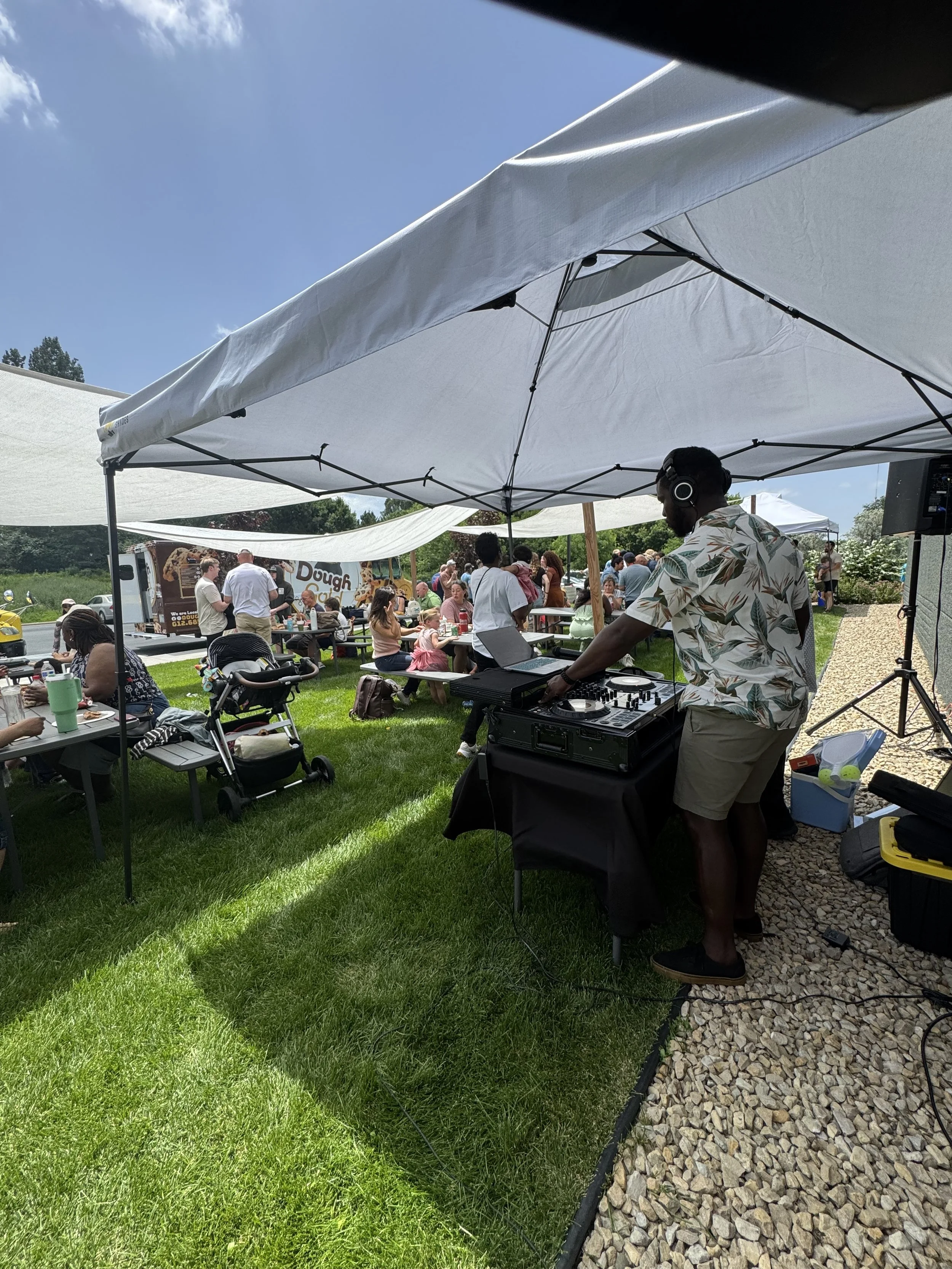 DJ playing music at an outdoor event with people sitting at tables and a food truck in the background.