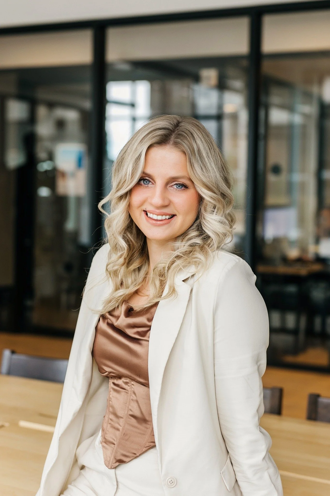 A smiling blonde woman in a white blazer, seated at a wooden table in a modern office with glass walls.