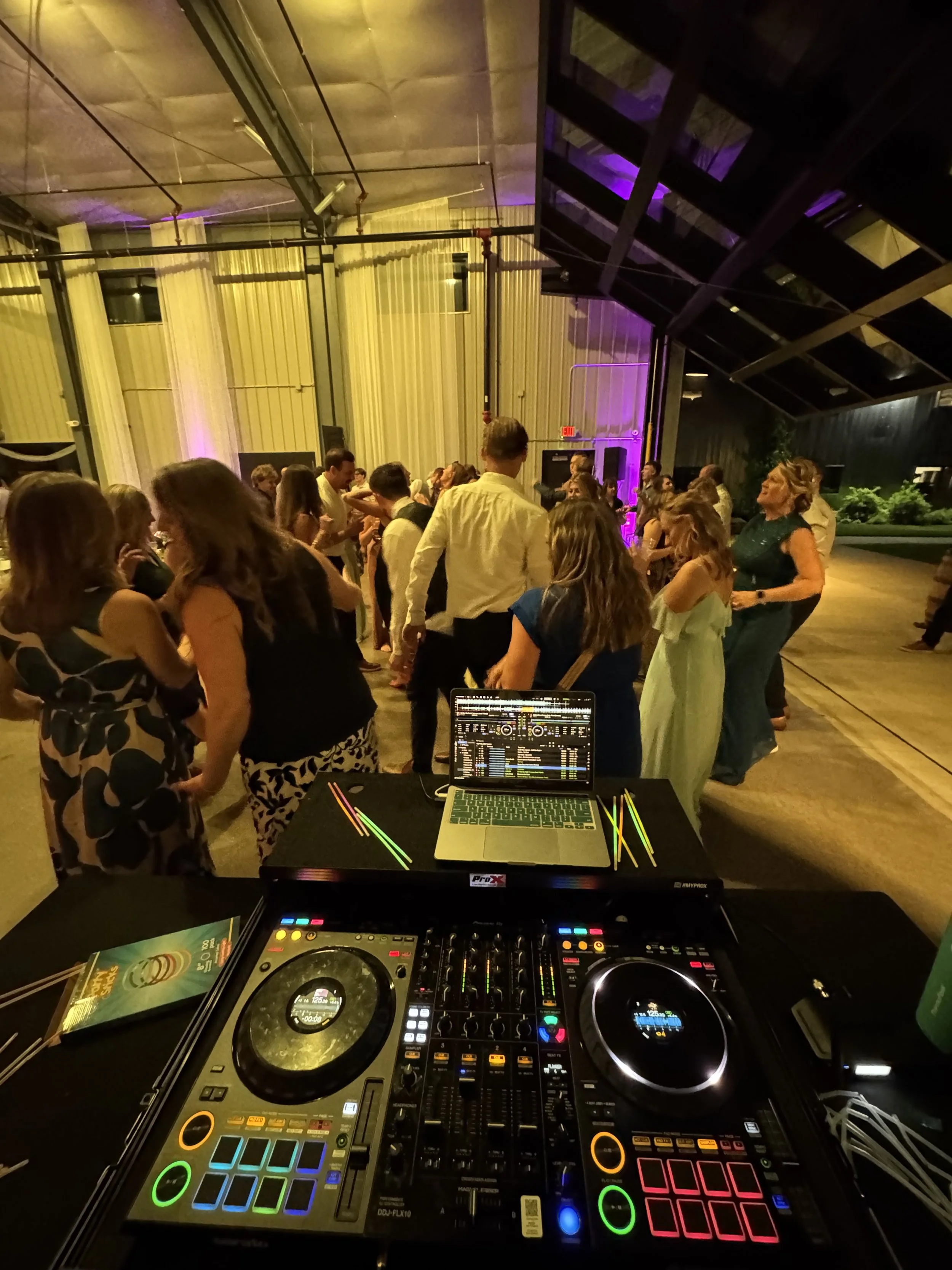 A DJ setup with turntables, a laptop with music software, and a booklet on a black table, overlooking a dance floor where people are dancing at an indoor party with purple lighting.