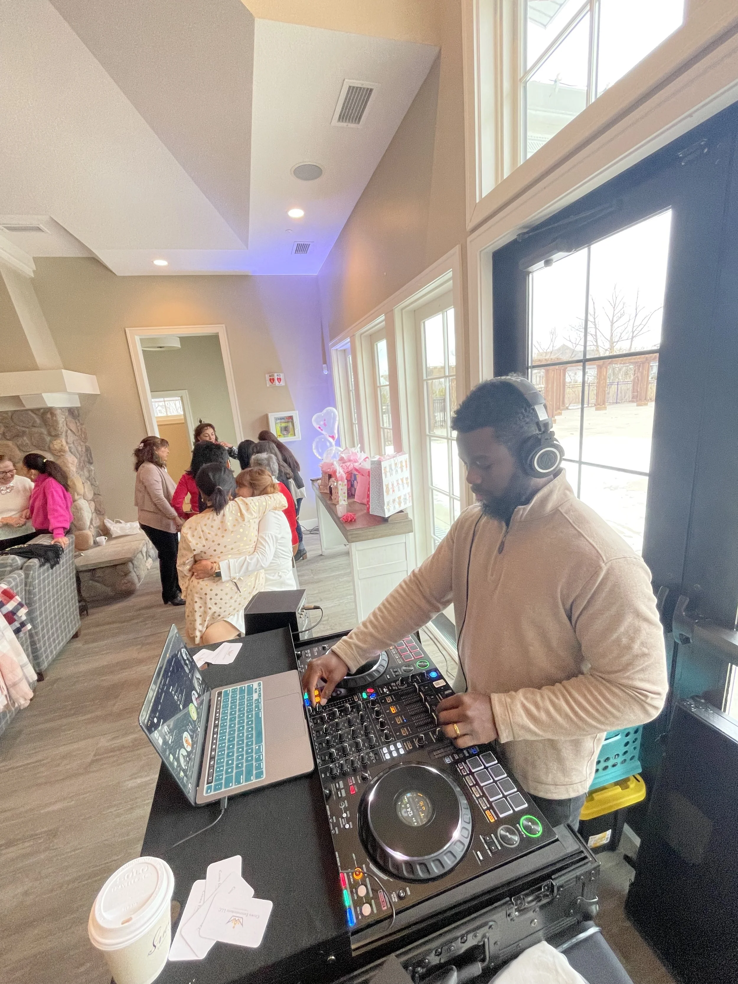 DJ playing music at a party with guests mingling and hugging indoors near a stone fireplace and large windows.