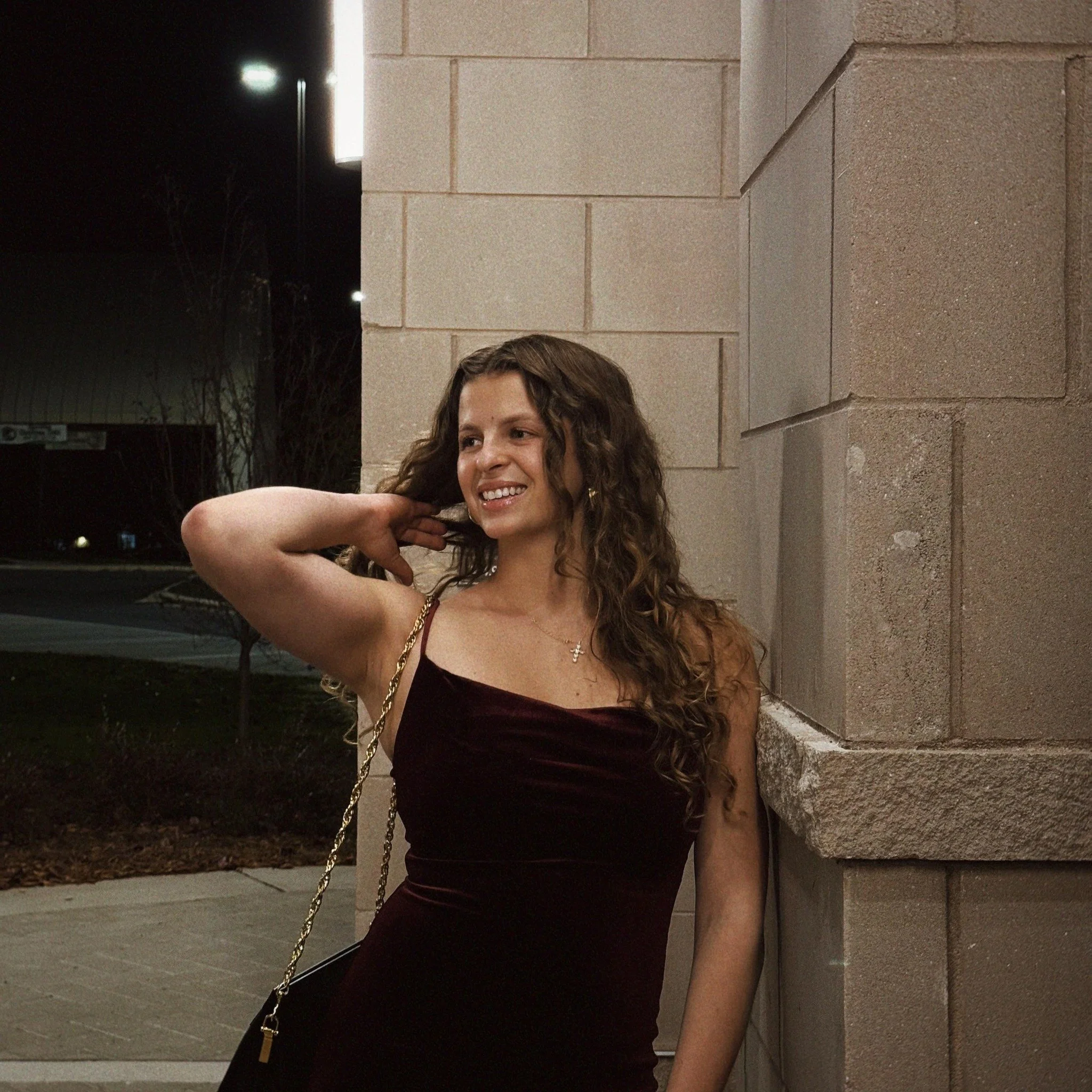 A woman with curly brown hair smiling and posing against a stone wall at night, wearing a burgundy dress and holding a small black purse with a gold chain.