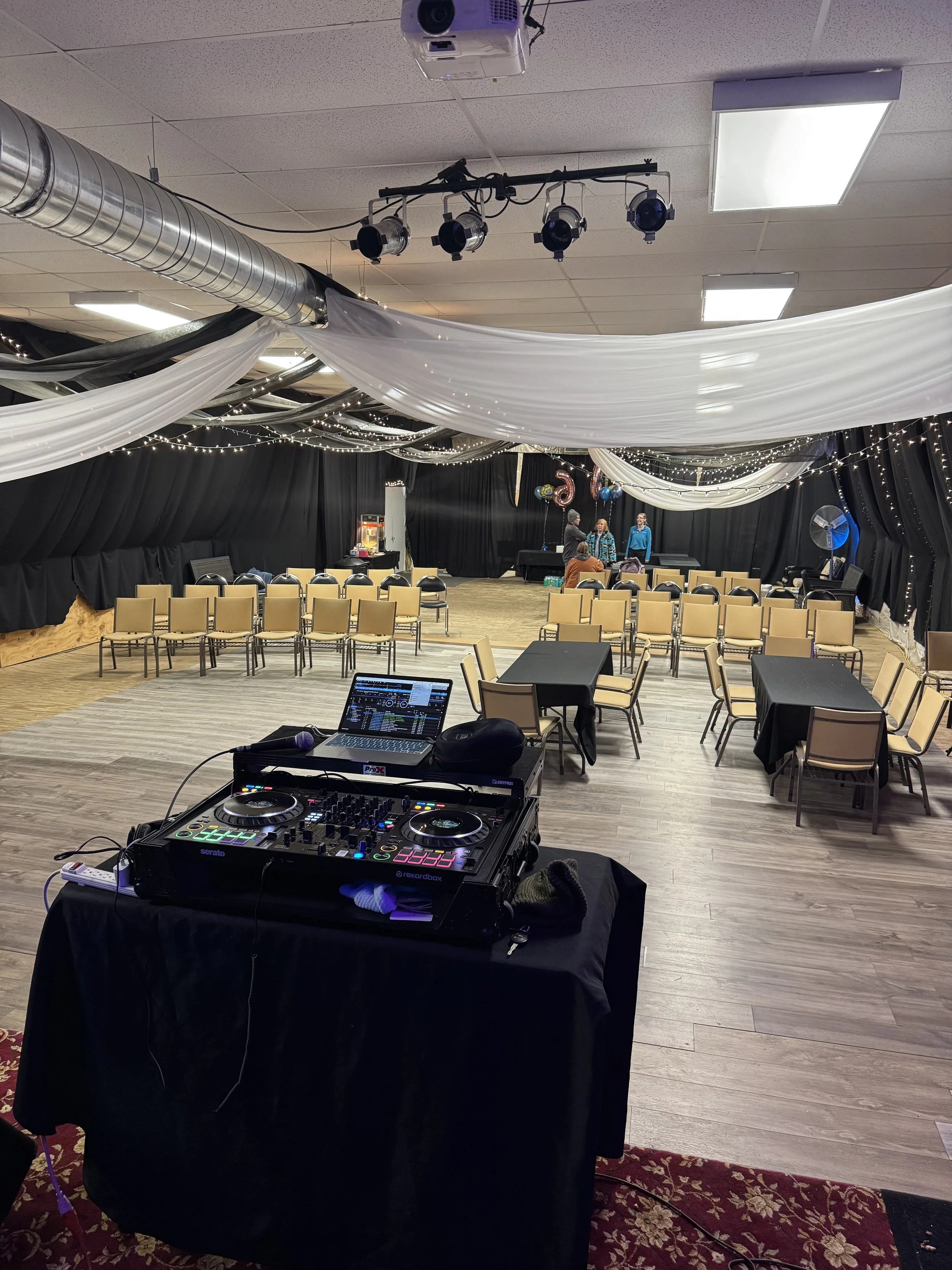 Event space with a DJ setup in the foreground, arranged tables and chairs, a stage with decorations, and ceiling drapes with string lights.
